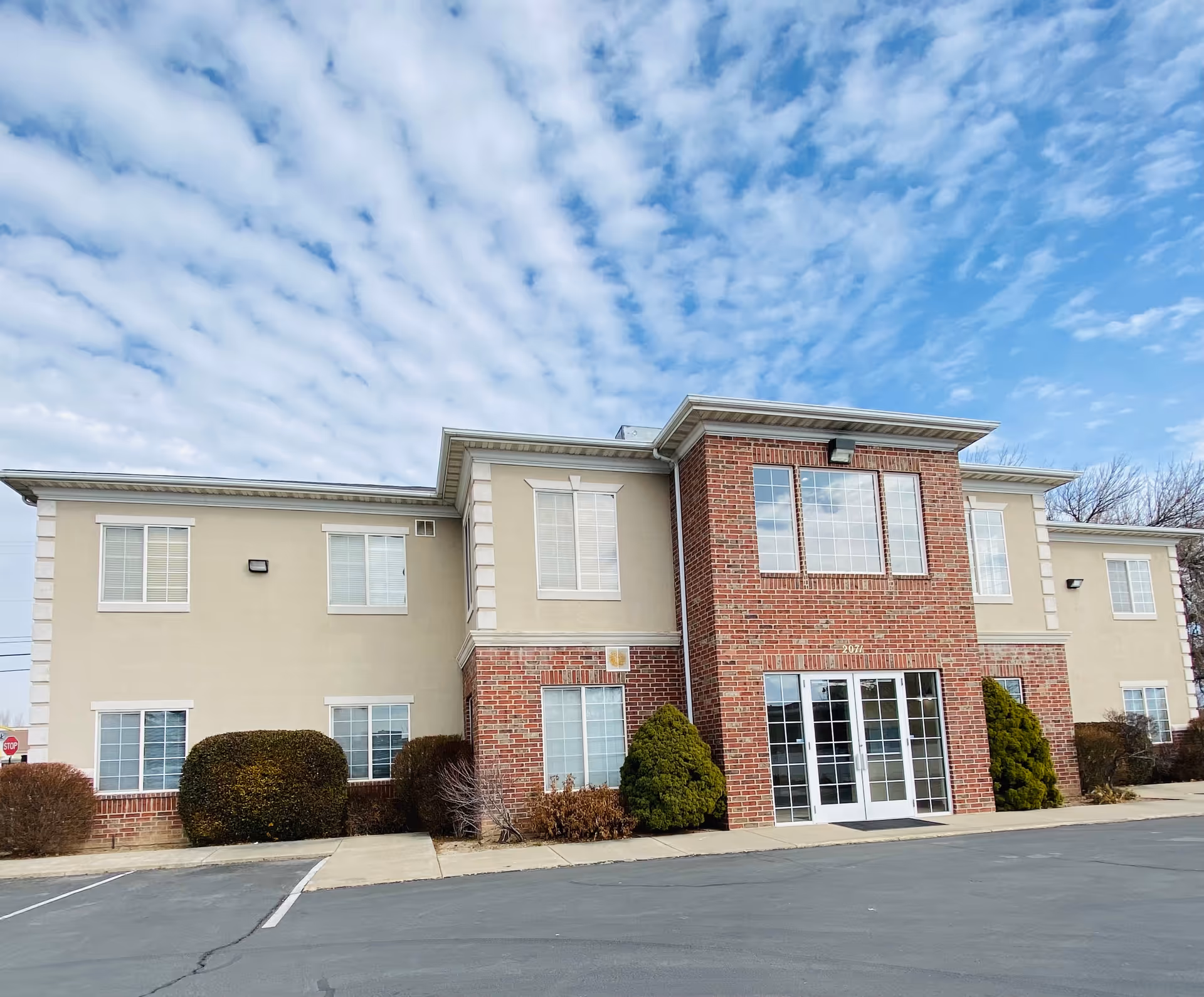 Two-story senior living building with brick entrance, large windows, and a parking lot in front.