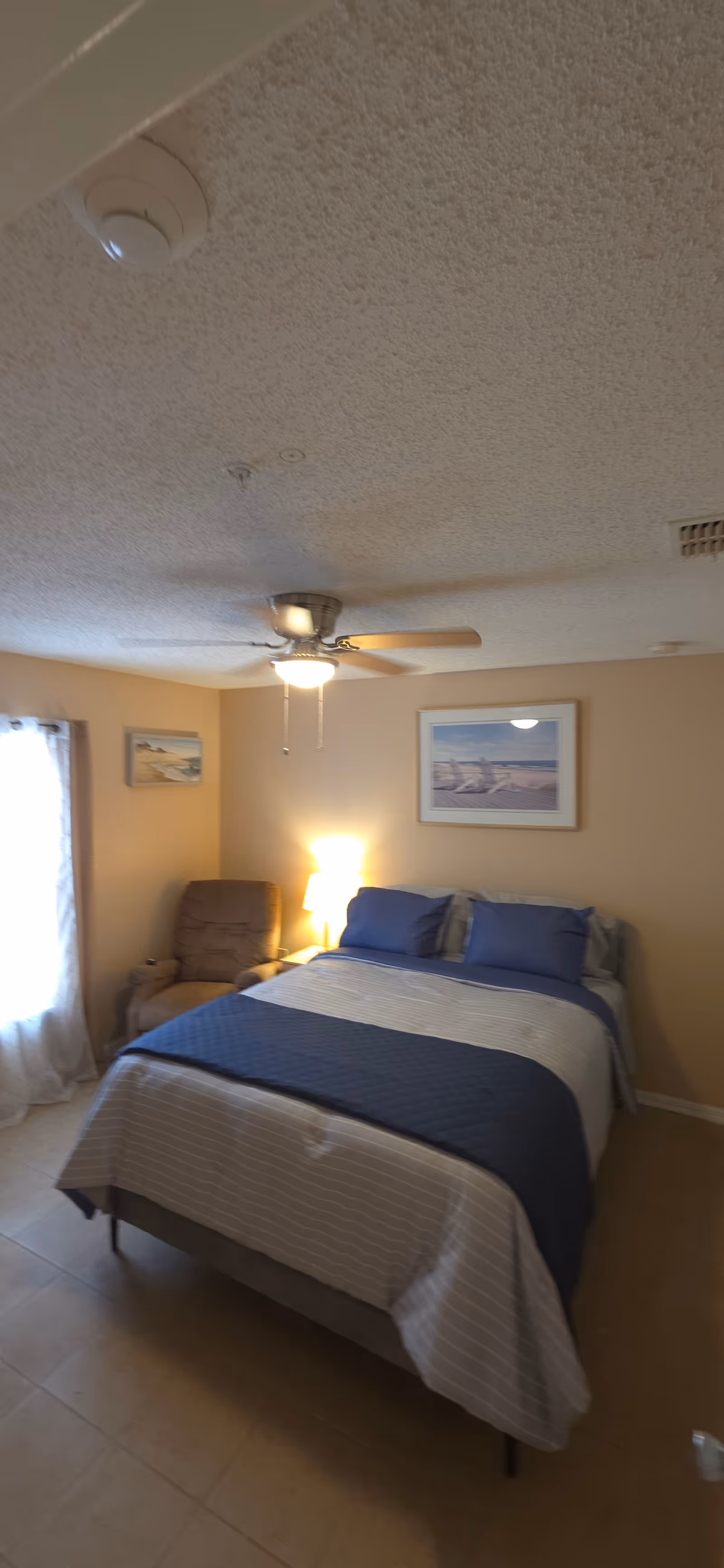 A cozy bedroom with a bed, blue bedding, chair, wall art, ceiling fan, and lamp.