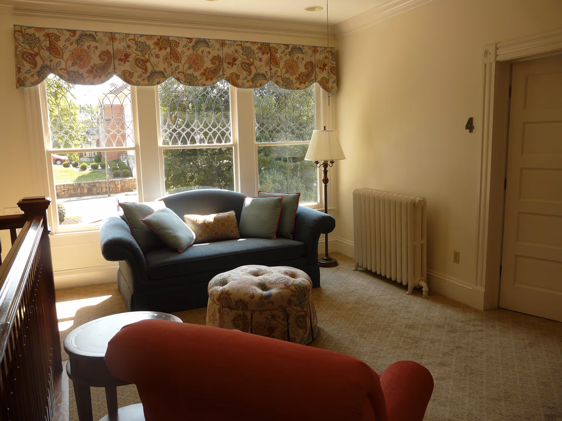 Cozy living room with blue sofa, patterned ottoman, large windows, and a standing lamp.
