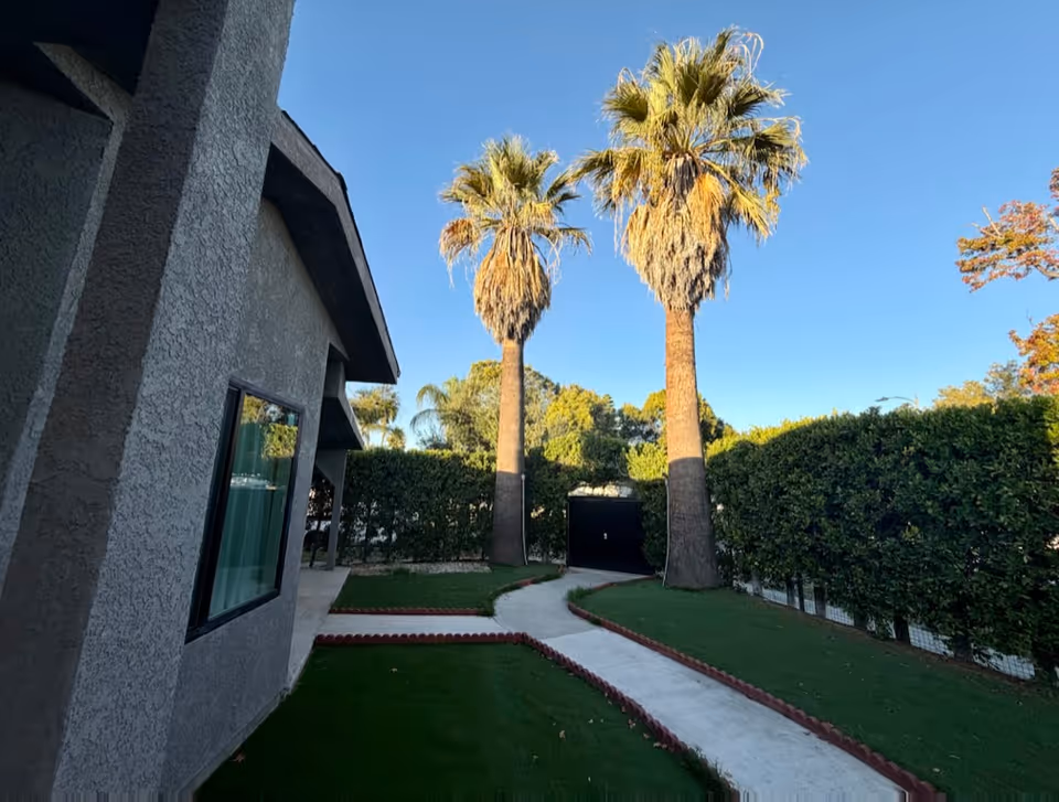 Exterior view of a gray stucco building with a winding concrete path, palm trees, and hedges.