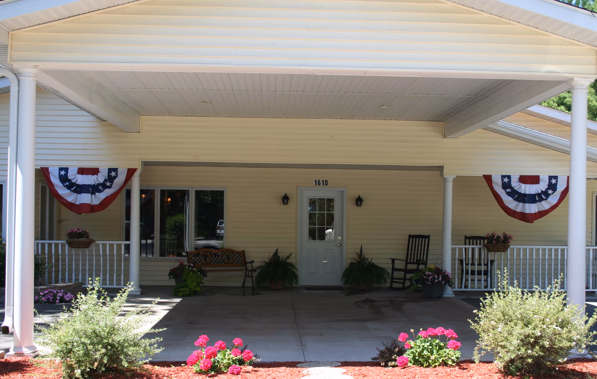 Covered front entrance with a door numbered 1610, a porch with rocking chairs, potted plants, and patriotic bunting.