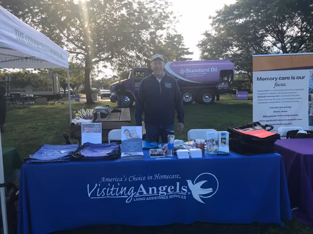 Man in cap stands behind a blue Visiting Angels booth at an outdoor event.