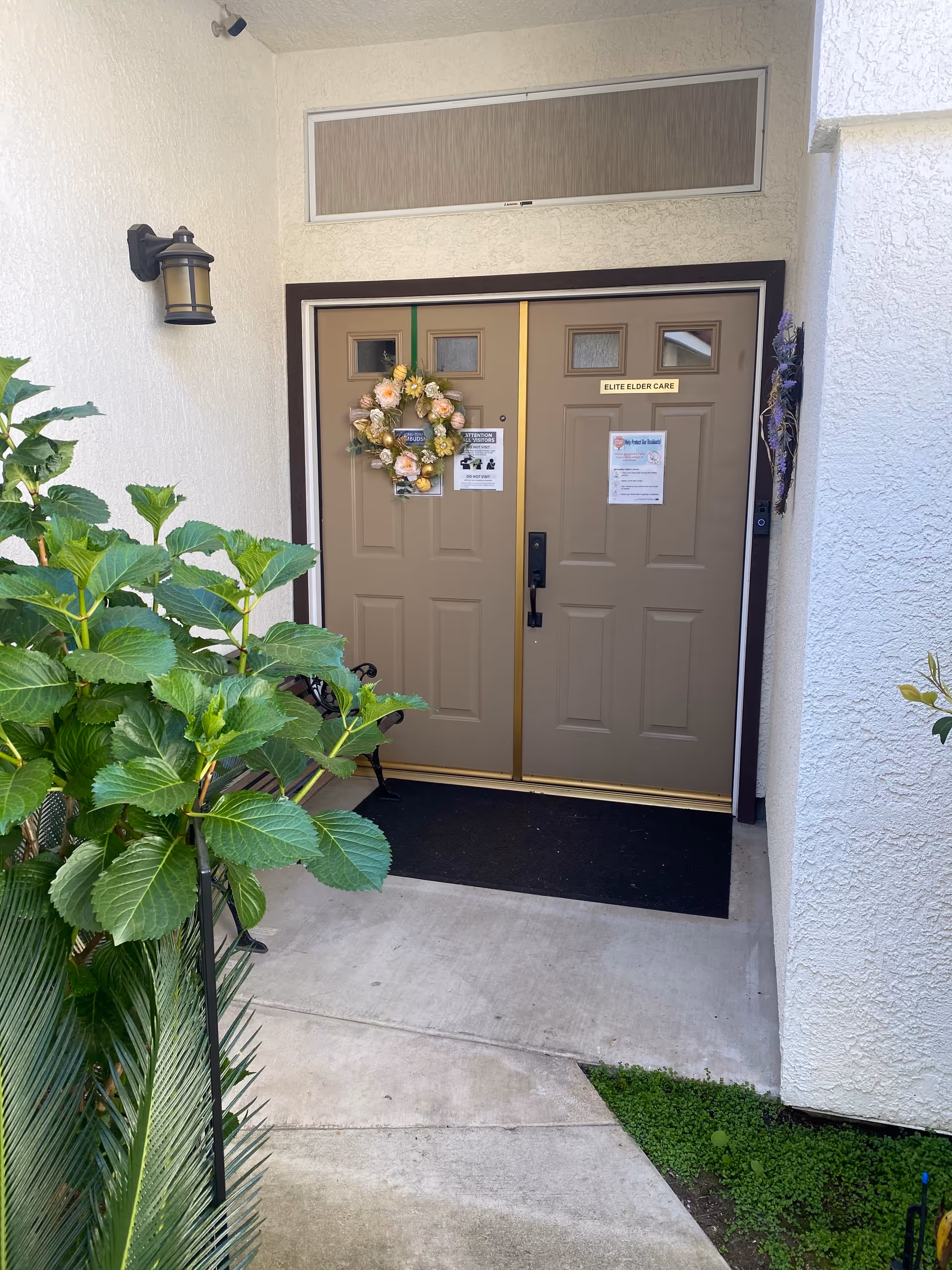 Double beige doors at a building entrance with a decorative wreath and surrounding greenery.