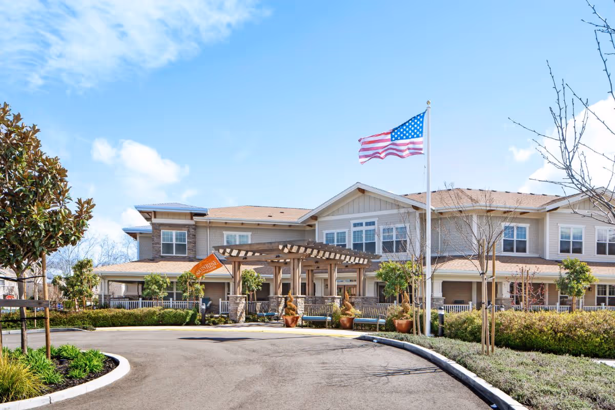 Exterior view of a senior living facility building with a curved driveway, landscaped greenery, an American flag on a flagpole, and a wooden pergola at the entrance under a clear blue sky.