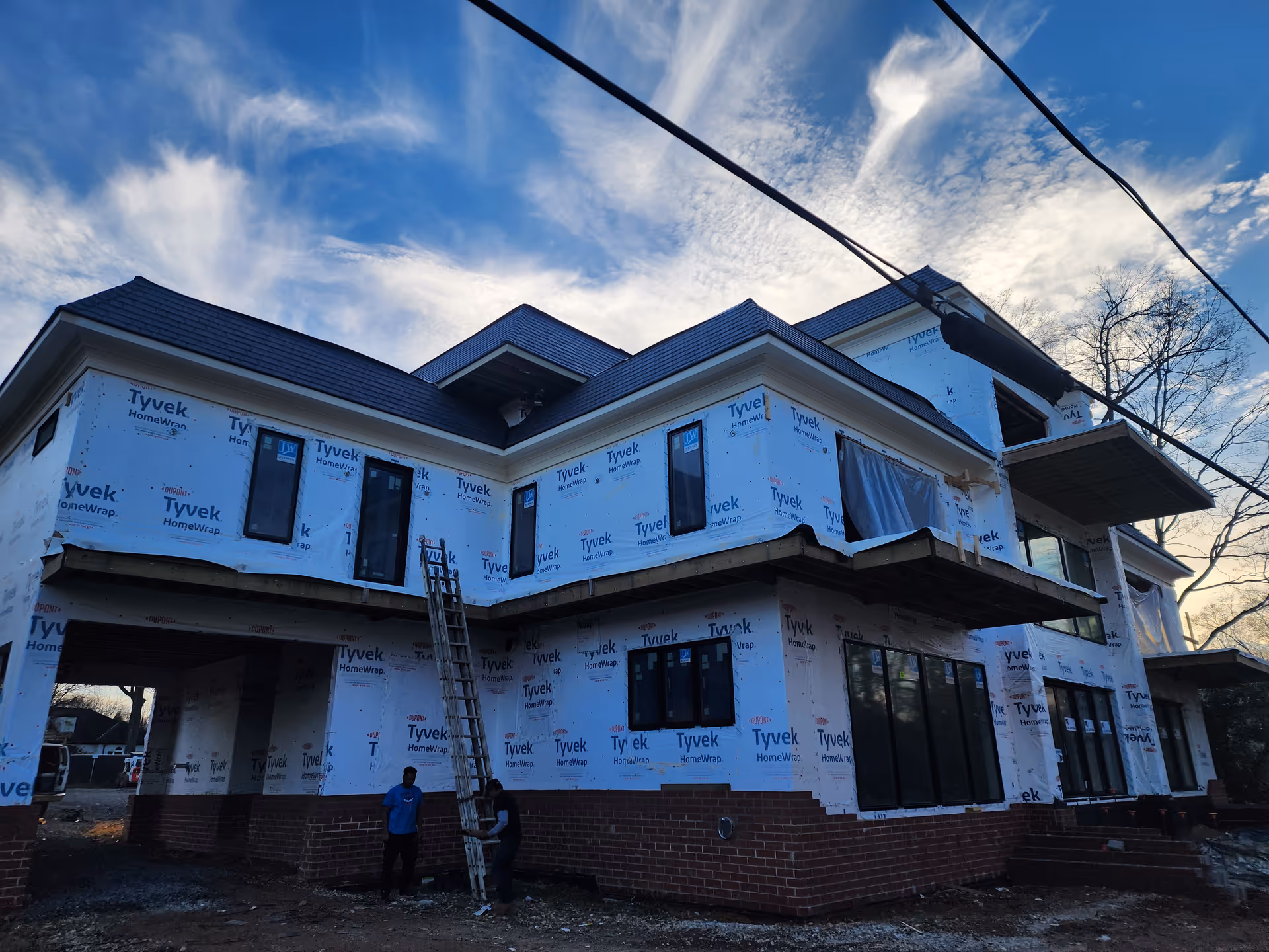Two-story building under construction wrapped in Tyvek with workers and a ladder outside.