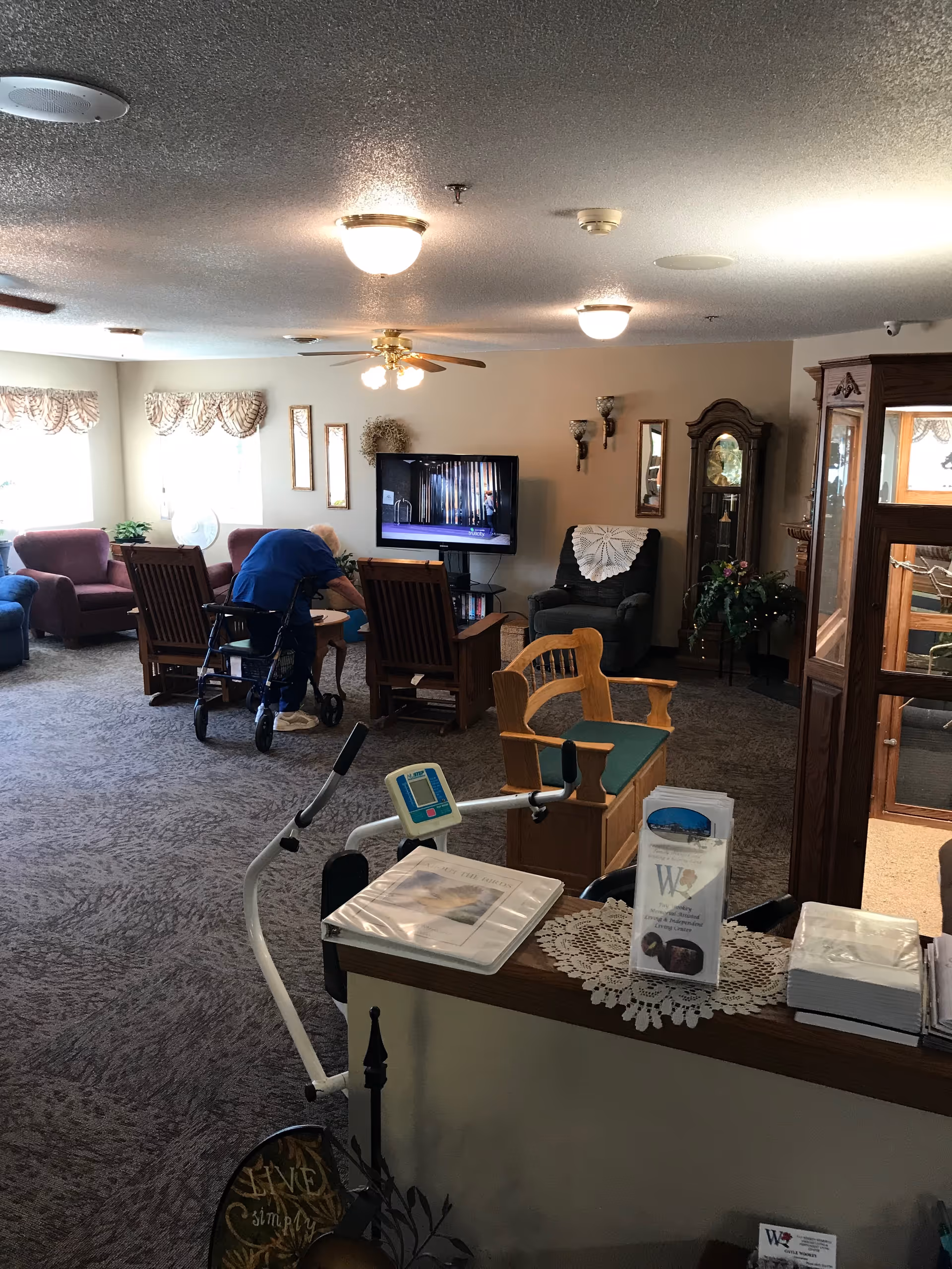 A cozy assisted living facility common area with carpeted floor, several armchairs, a television mounted on the wall, a grandfather clock, and a person using a walker bending over near a table. There is a wooden bench with green cushions, a small exercise machine, and a counter with brochures and a doily in the foreground.