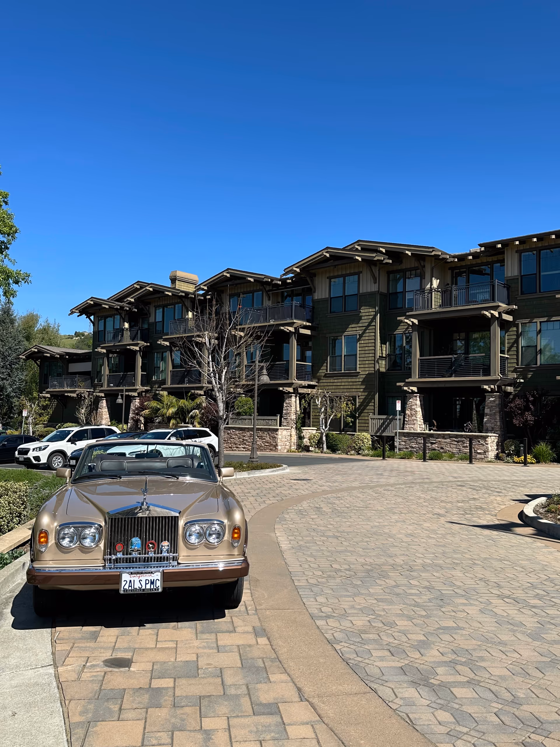 A tan vintage car is parked in front of a multi-story senior living building.