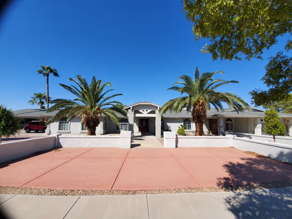 Front exterior view of a single-story adult care home building with a wide driveway, two large palm trees in front, and a clear blue sky overhead.