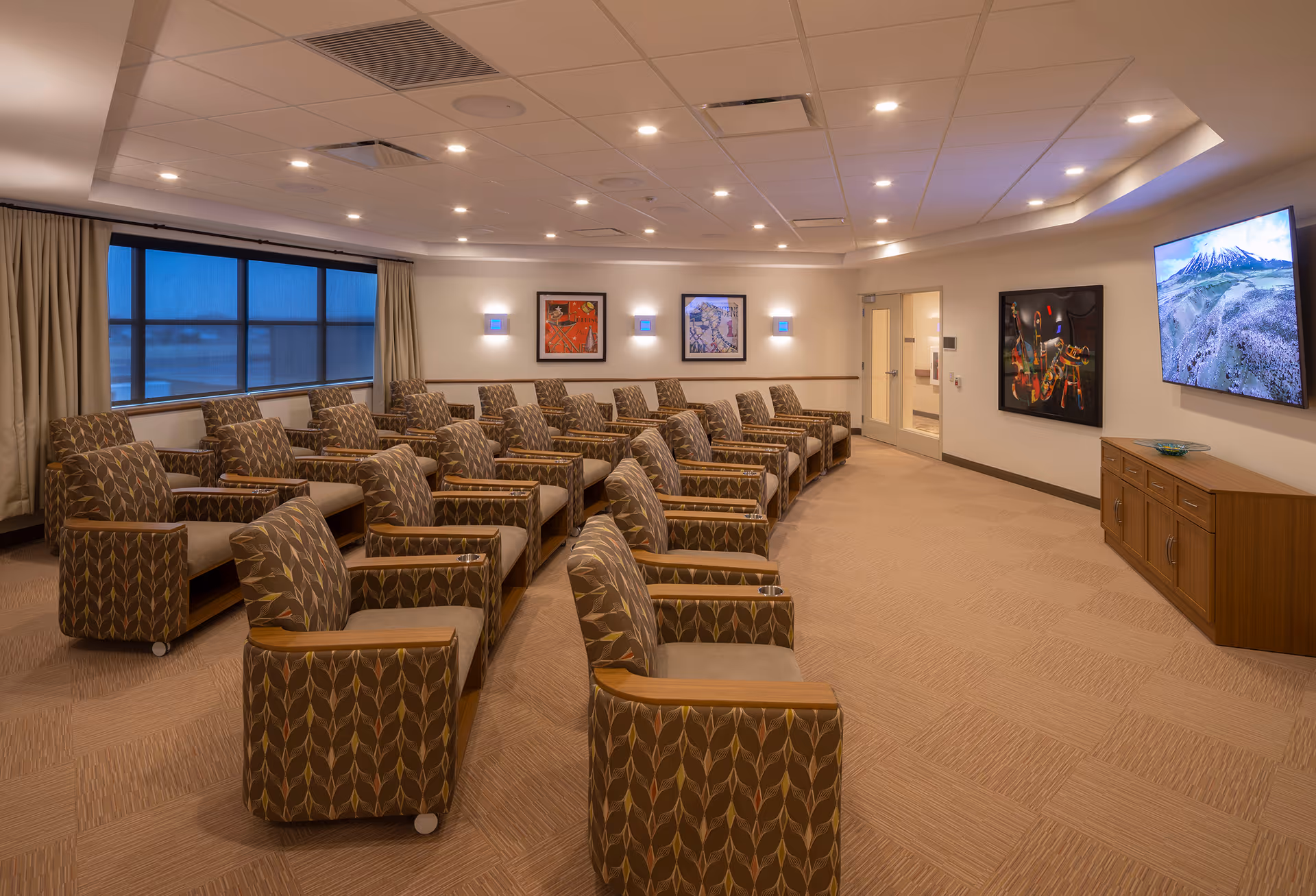 Rows of patterned chairs face a wall-mounted TV in a spacious senior living lounge.