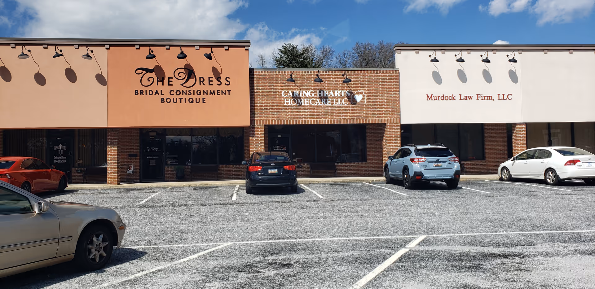 Strip mall exterior featuring Caring Hearts Homecare LLC sign between other storefronts and parked cars.