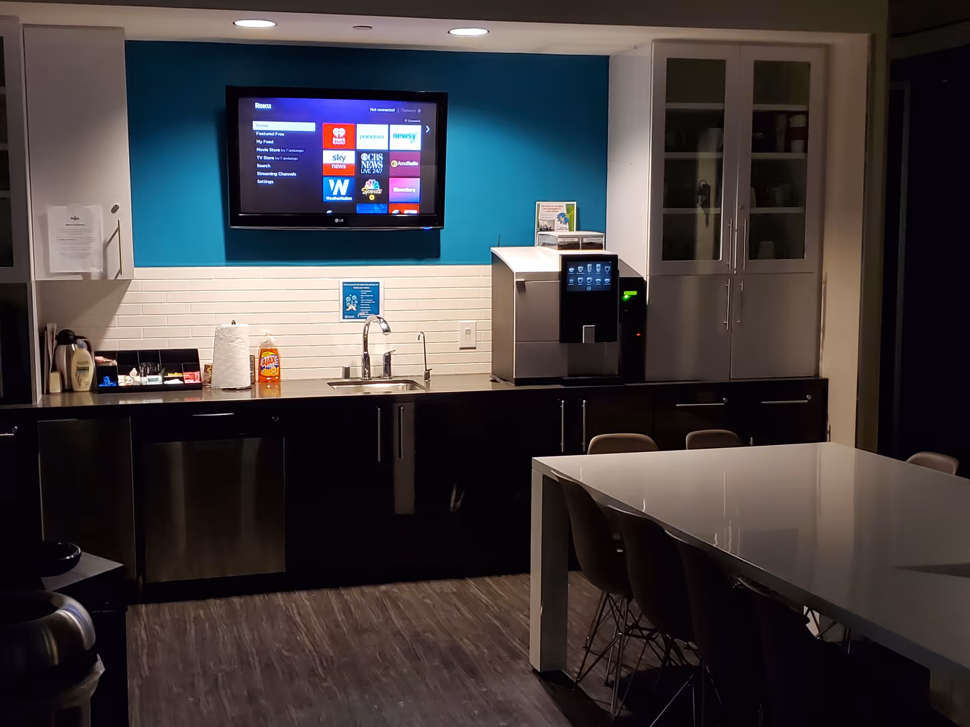 Community kitchenette with teal wall, TV, coffee machine, sink, and long dining table.