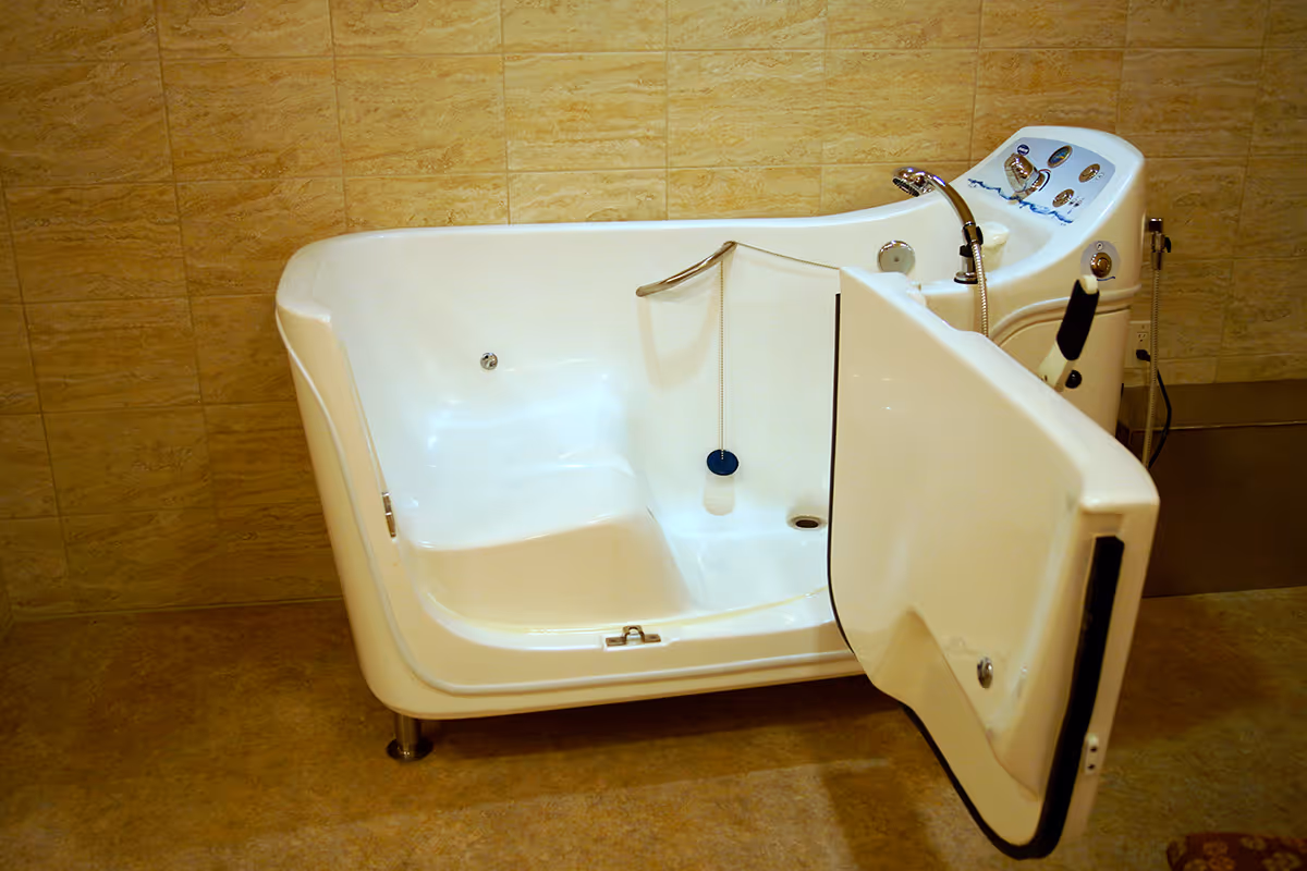 A white walk-in bathtub with an open door, designed for easy access, situated against a beige tiled wall and floor. The bathtub has built-in controls and a handheld showerhead on the right side.