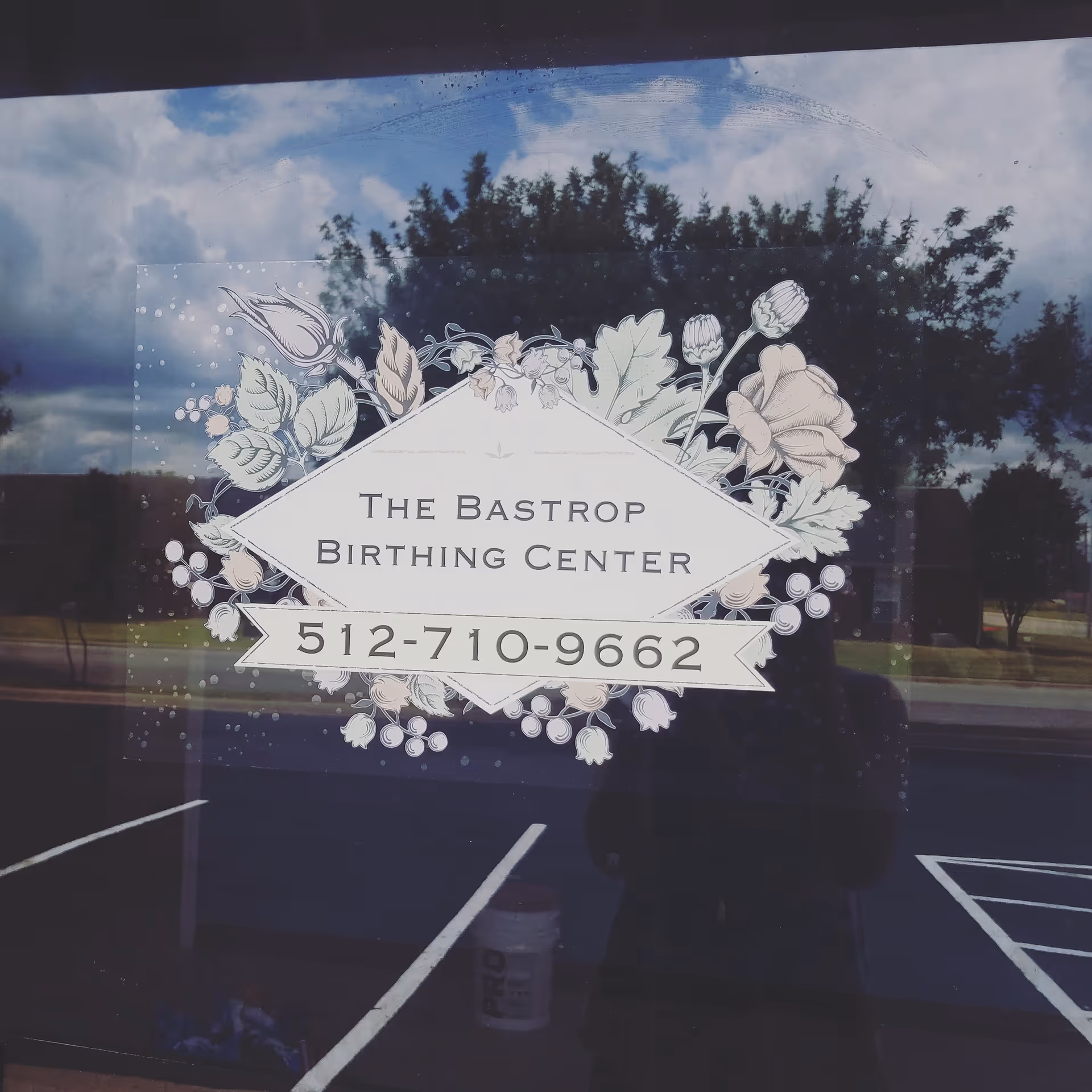 Glass storefront window displaying The Bastrop Birthing Center sign with floral border and reflections.