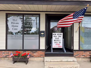 Front of TLC Private Home Care building featuring flag, hiring sign, and flowers.