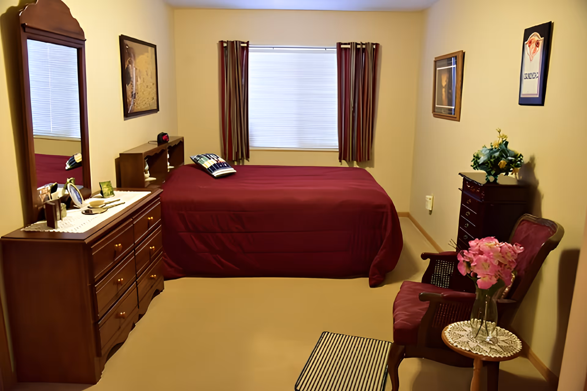 Cozy bedroom with a burgundy-covered bed, wooden dresser and mirror, armchair with flowers, and a window with striped curtains.