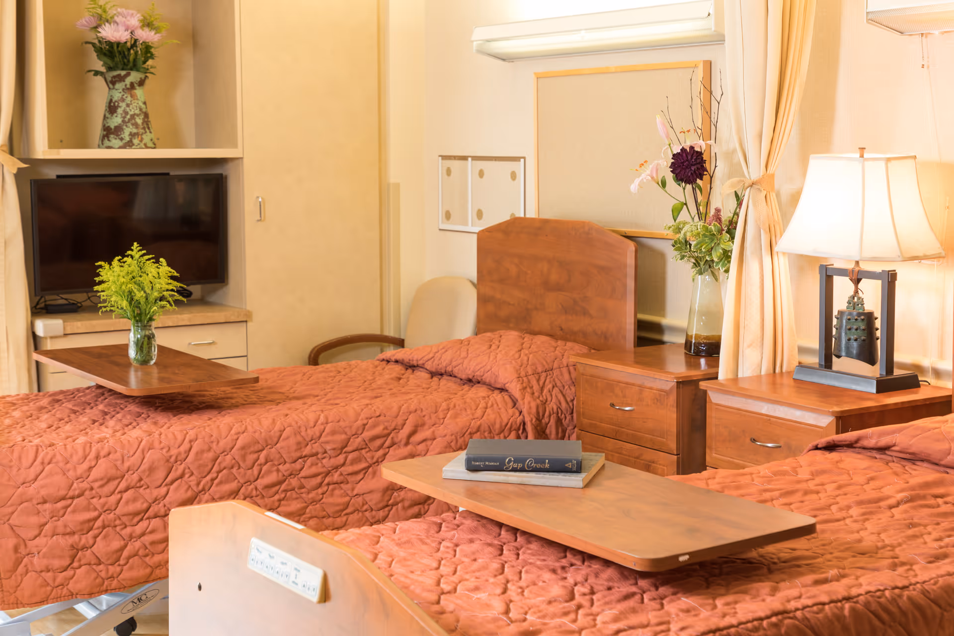 Two single beds with wooden headboards and rust-colored quilts in a well-lit senior living bedroom with bedside tables, lamps, and floral arrangements.