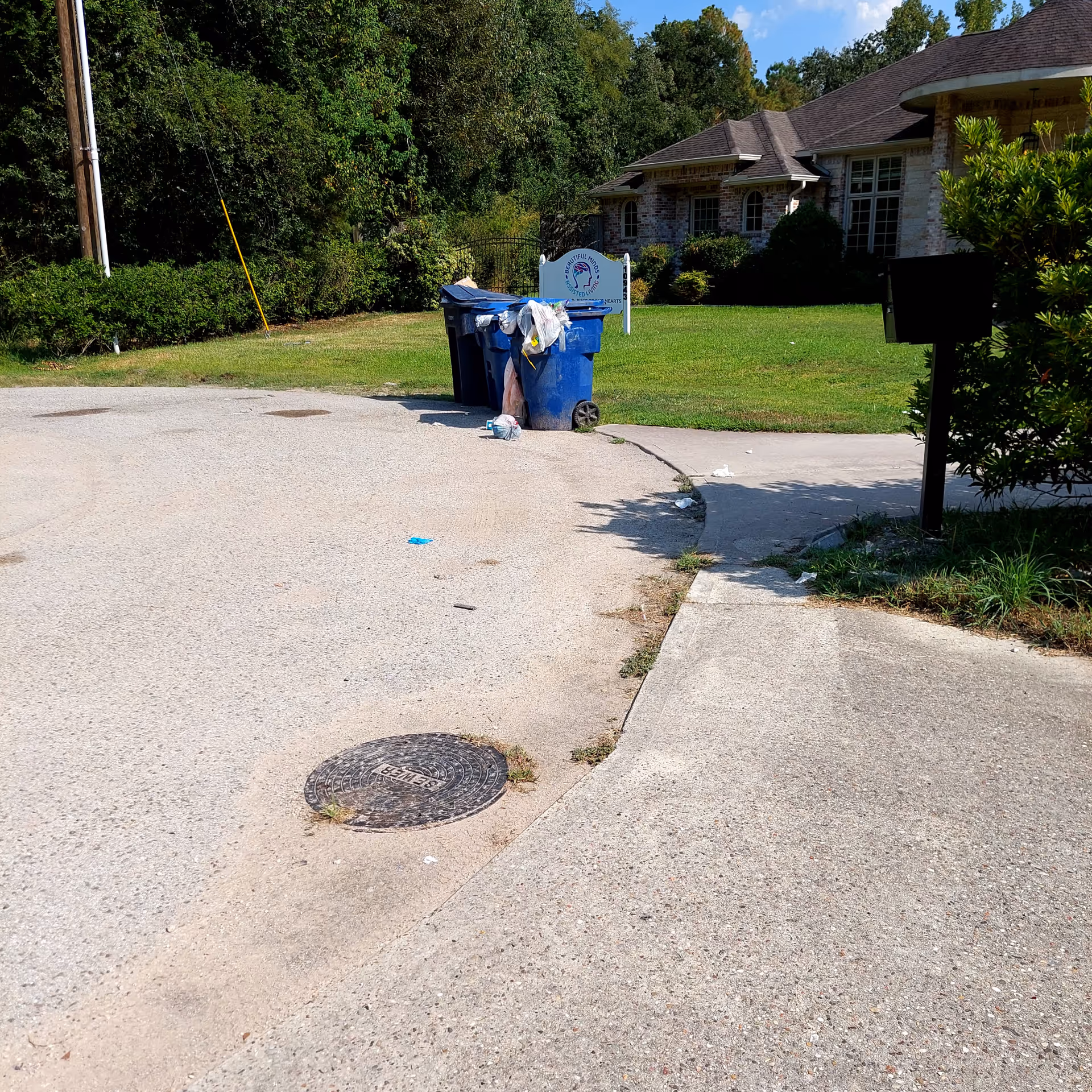 A suburban curb with blue trash bins in front of a brick house and green yard.