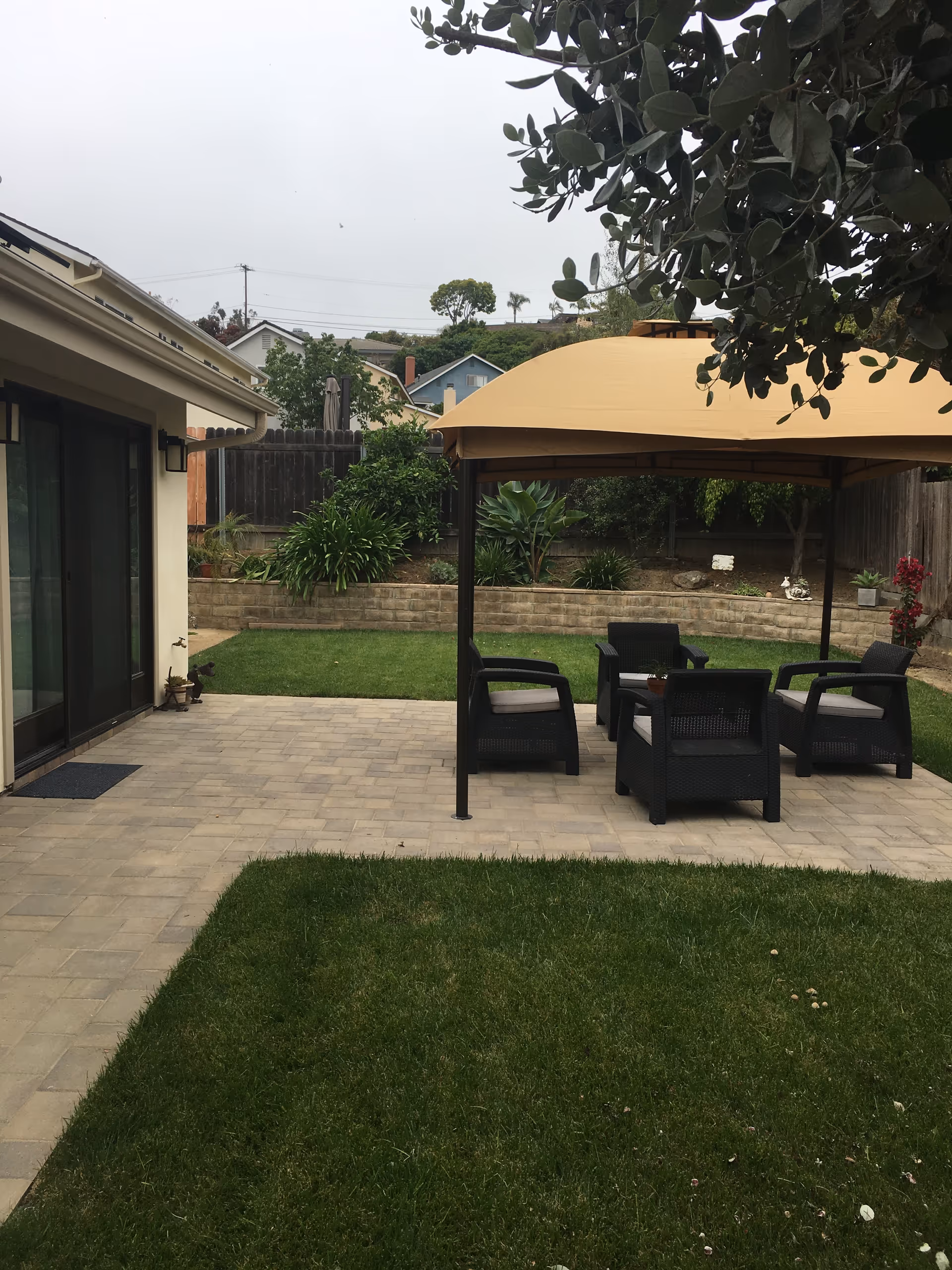 Outdoor patio area with a beige canopy covering four black chairs arranged around a small table. The patio is paved with light-colored bricks and is adjacent to a grassy lawn. There are plants and shrubs along a raised garden bed with a stone retaining wall in the background, and a wooden fence behind the garden bed. Part of a house with sliding glass doors is visible on the left side of the image.