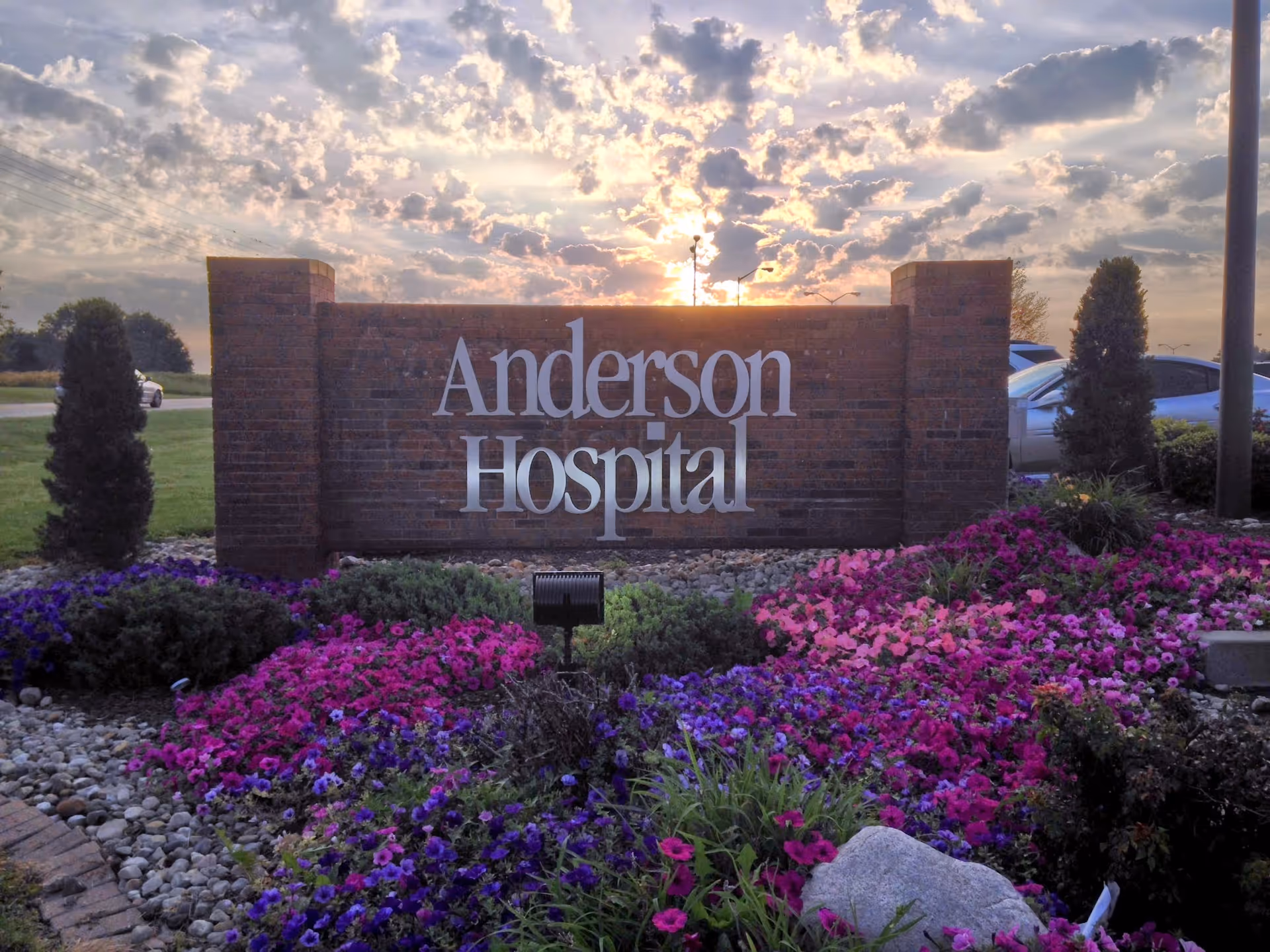 Brick hospital sign reading Anderson Hospital surrounded by colorful flowers under a sunset sky