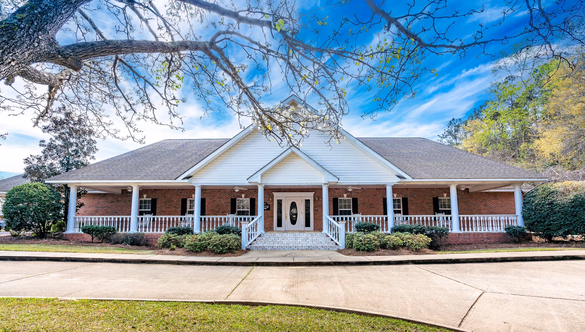 Front view of a single-story brick retirement home with a covered wraparound porch, white columns, entrance steps and a driveway, framed by tree branches.