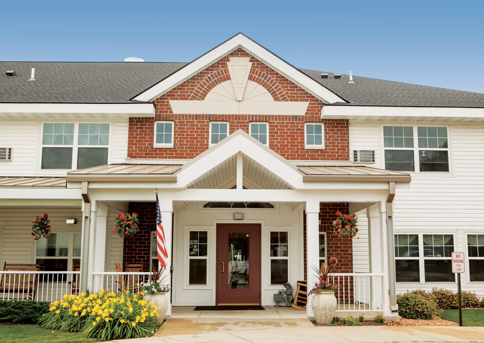 Front exterior view of a senior living facility building with a covered entrance, brick and white siding facade, hanging flower baskets, an American flag, and yellow flowers in the landscaping.