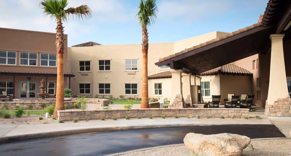 Exterior view of an assisted living building with a covered entrance, palm trees, and a courtyard with seating.