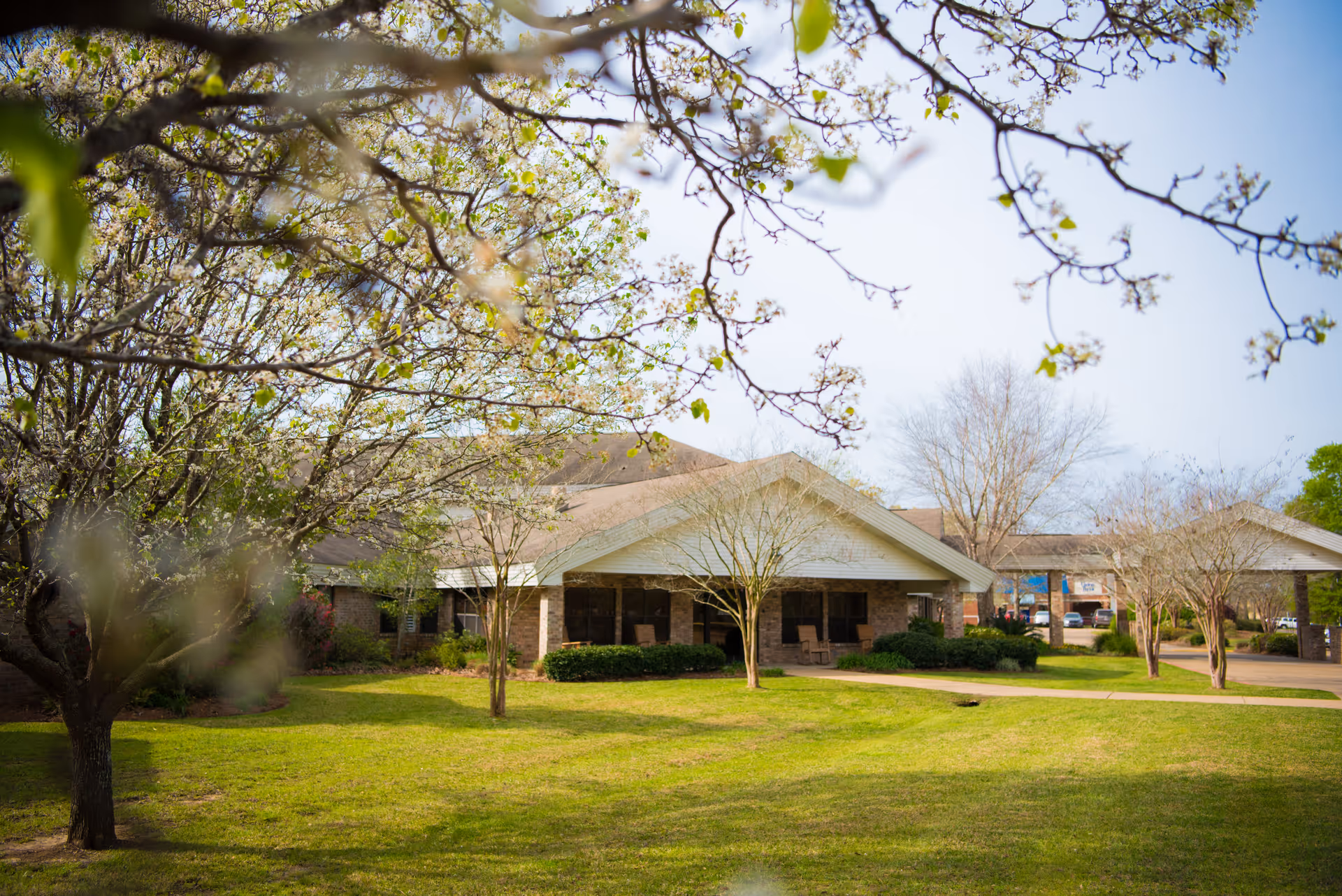 View of a single-story brick building with a covered porch and several rocking chairs, surrounded by a green lawn and trees with budding leaves under a clear sky.