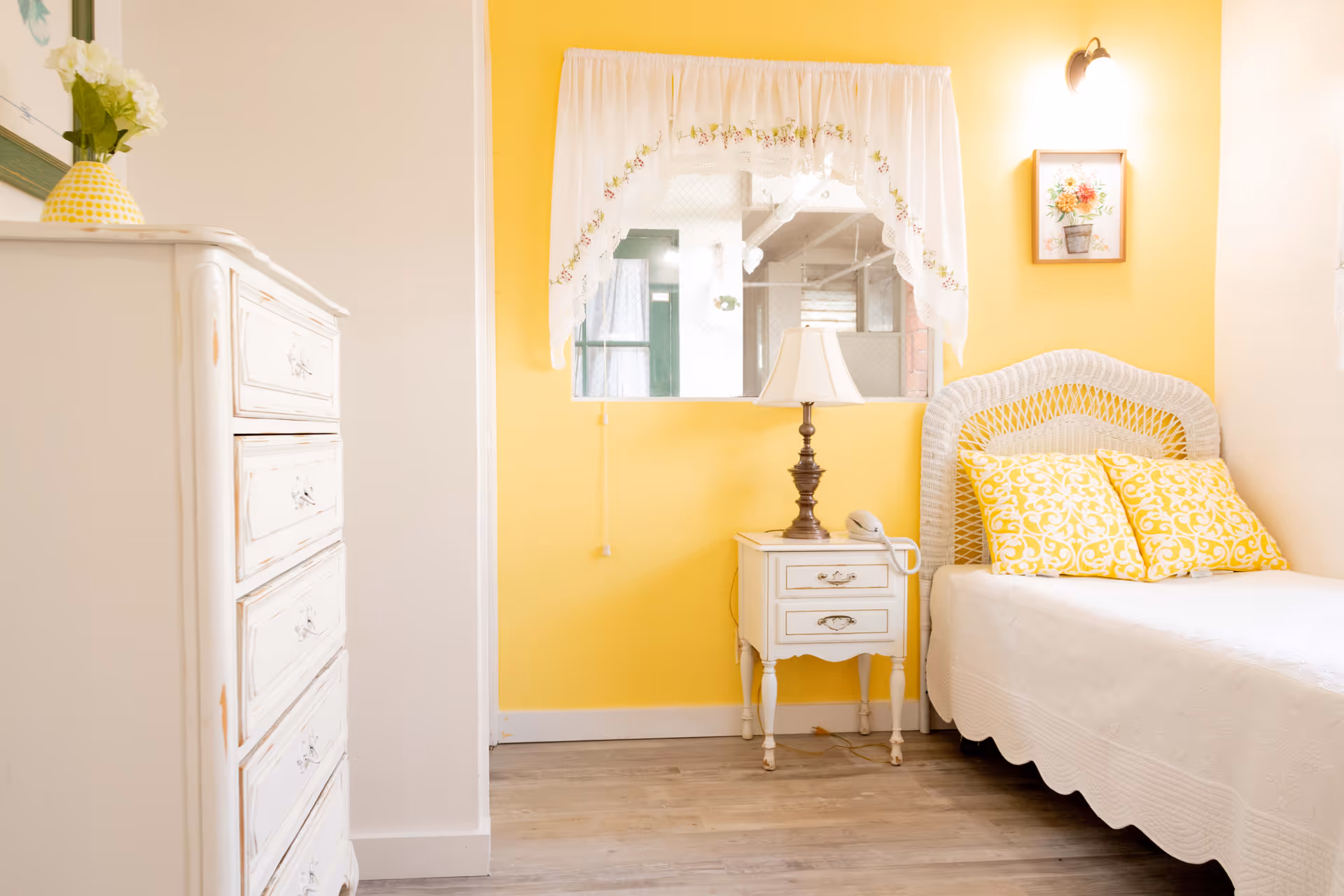 Yellow-walled bedroom with white furniture, wicker headboard, and lamp.