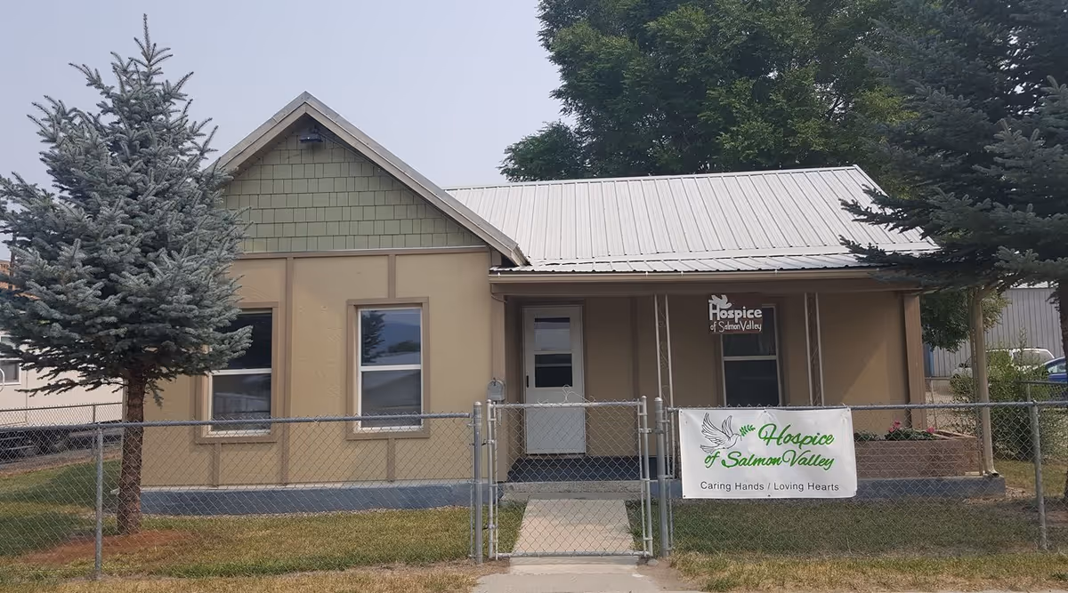 Beige building with metal roof, front entrance, and chain-link fence banner for Hospice of Salmon Valley.