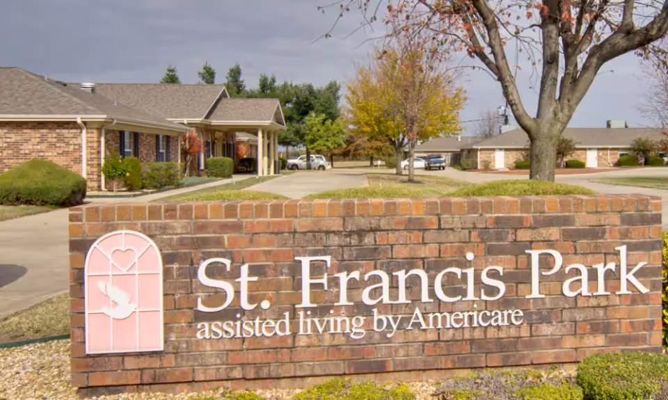 Brick sign for St. Francis Park assisted living by Americare in front of a driveway and single-story brick buildings with trees and shrubs around.