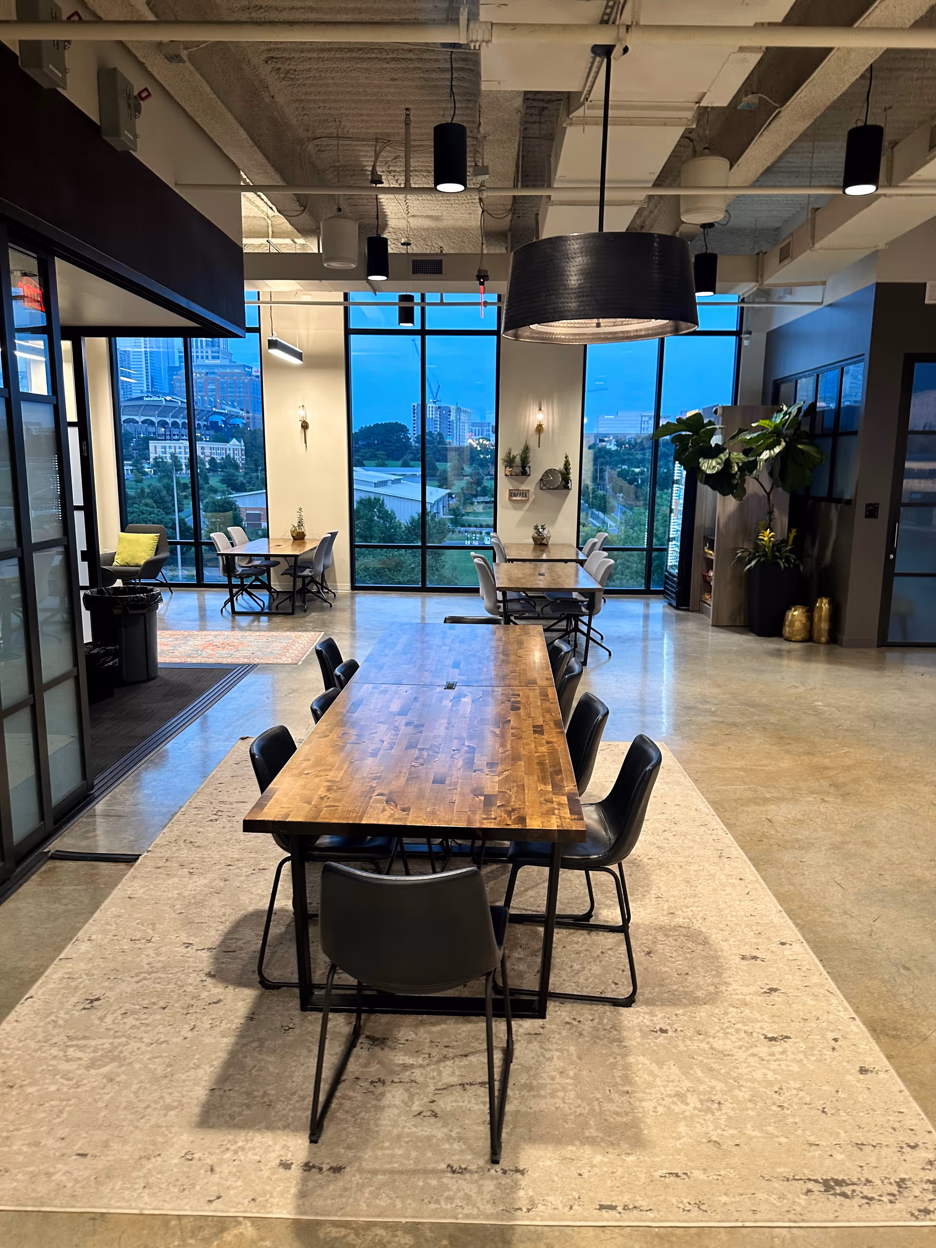 Modern communal dining area with long wooden tables and black chairs by large windows.