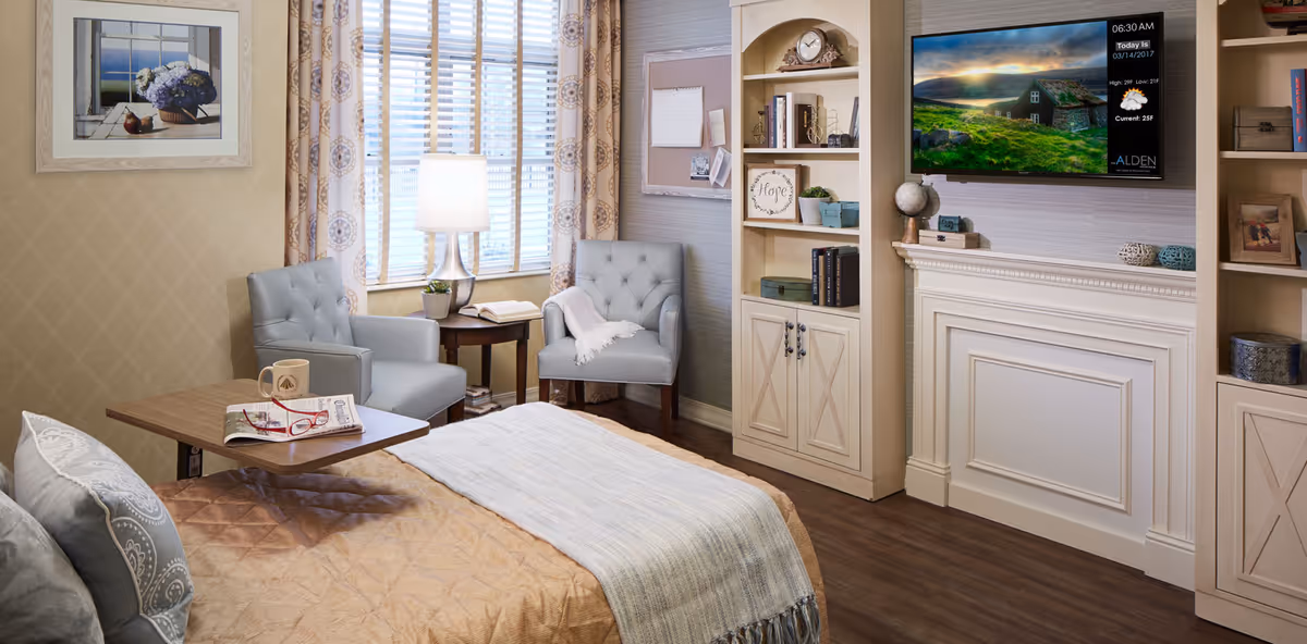 A cozy senior living room with a bed covered in a tan quilt and a light blue throw blanket. Two light blue armchairs are positioned near a window with patterned curtains and a table lamp. A small wooden table holds a coffee mug, a newspaper, and red reading glasses. Built-in shelves and cabinets flank a white fireplace mantel with decorative items and a mounted flat-screen TV displaying weather information and the Alden Estates logo.