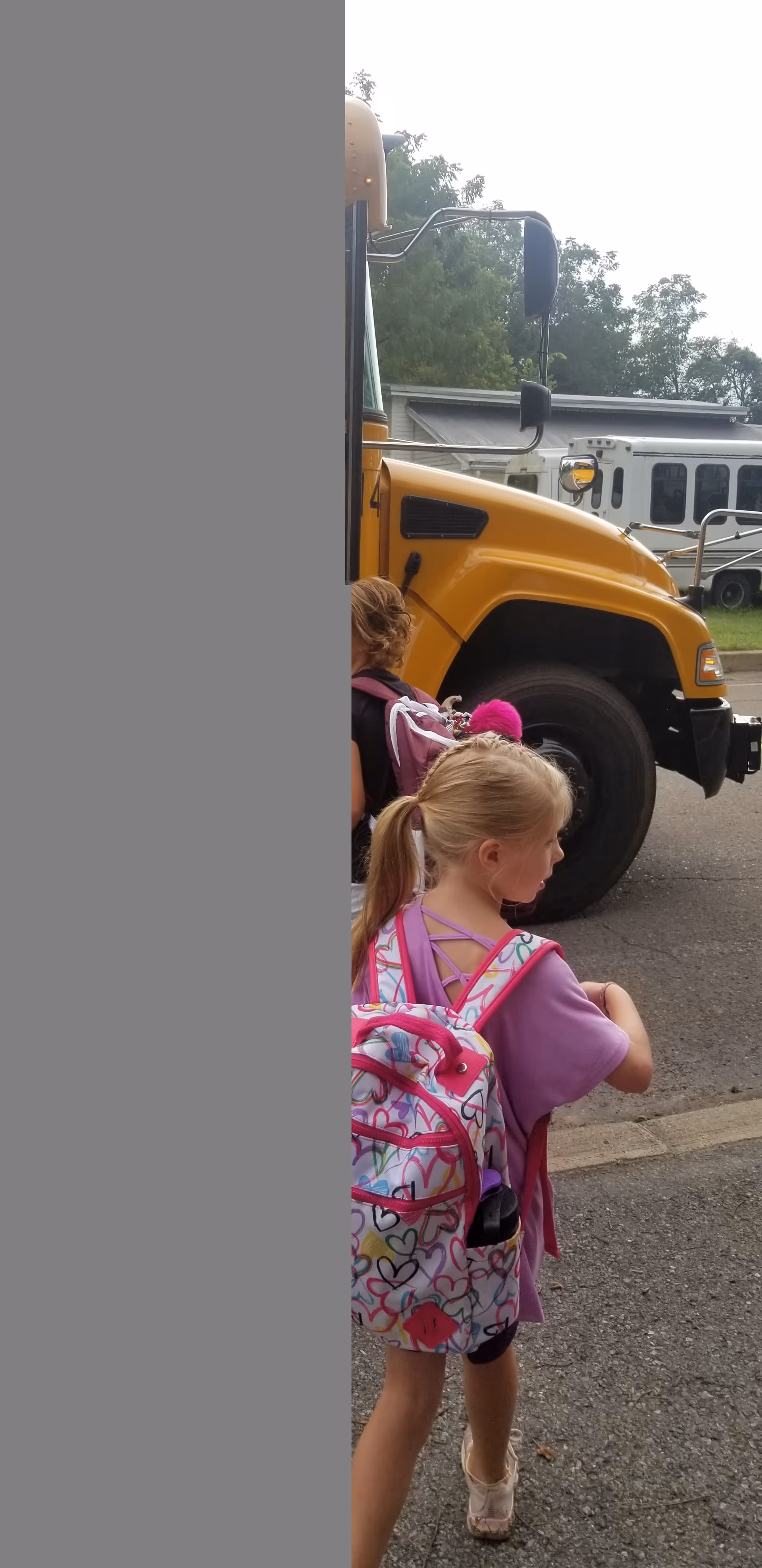 A young girl with a pink backpack stands near a yellow school bus outdoors.