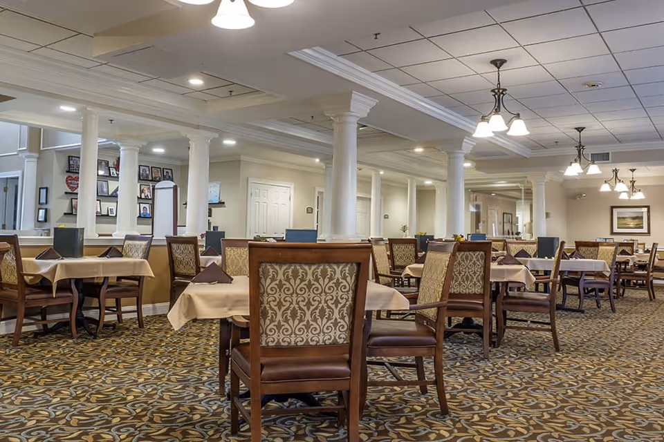 A spacious dining room in a senior living facility with multiple tables covered with beige tablecloths and set with folded brown napkins. The room features patterned carpet, white columns, and ceiling lights with multiple bulbs. There are framed pictures on the walls and a calm, inviting atmosphere.