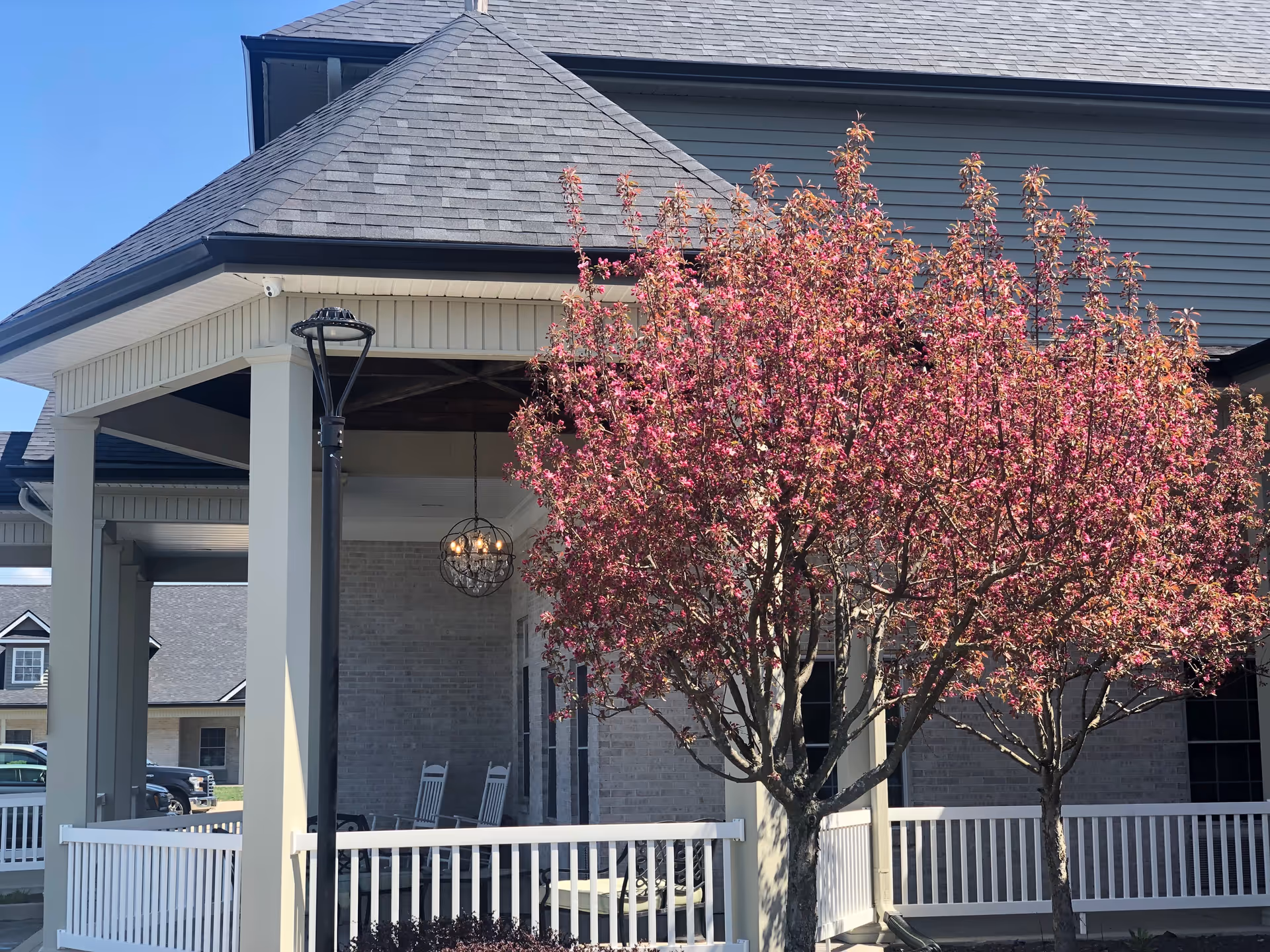 Front entrance of The Oaks at Plainfield with a covered porch, flowering pink trees, and railing.