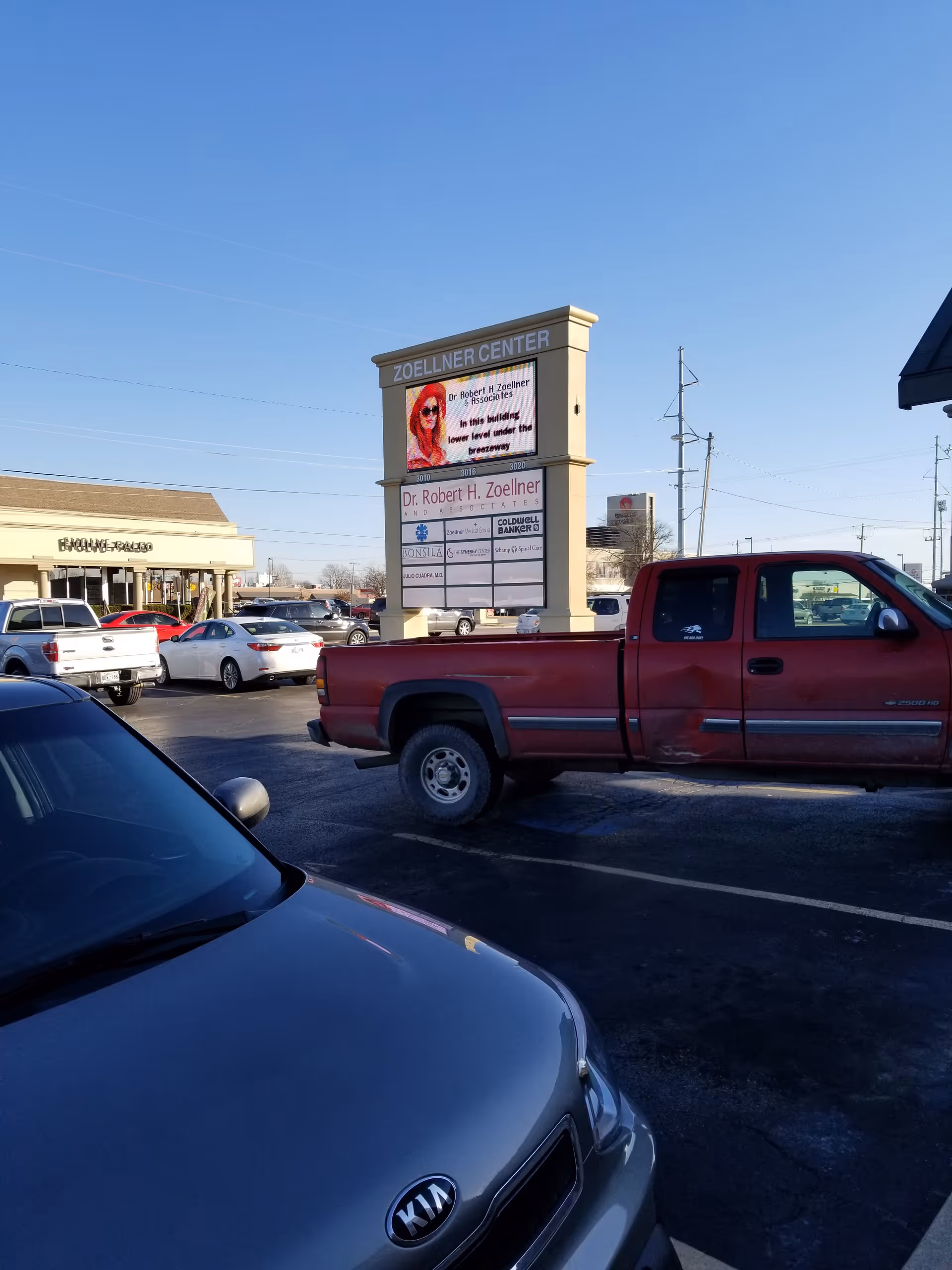 A parking lot with several cars and a tall signboard in front of a strip mall.