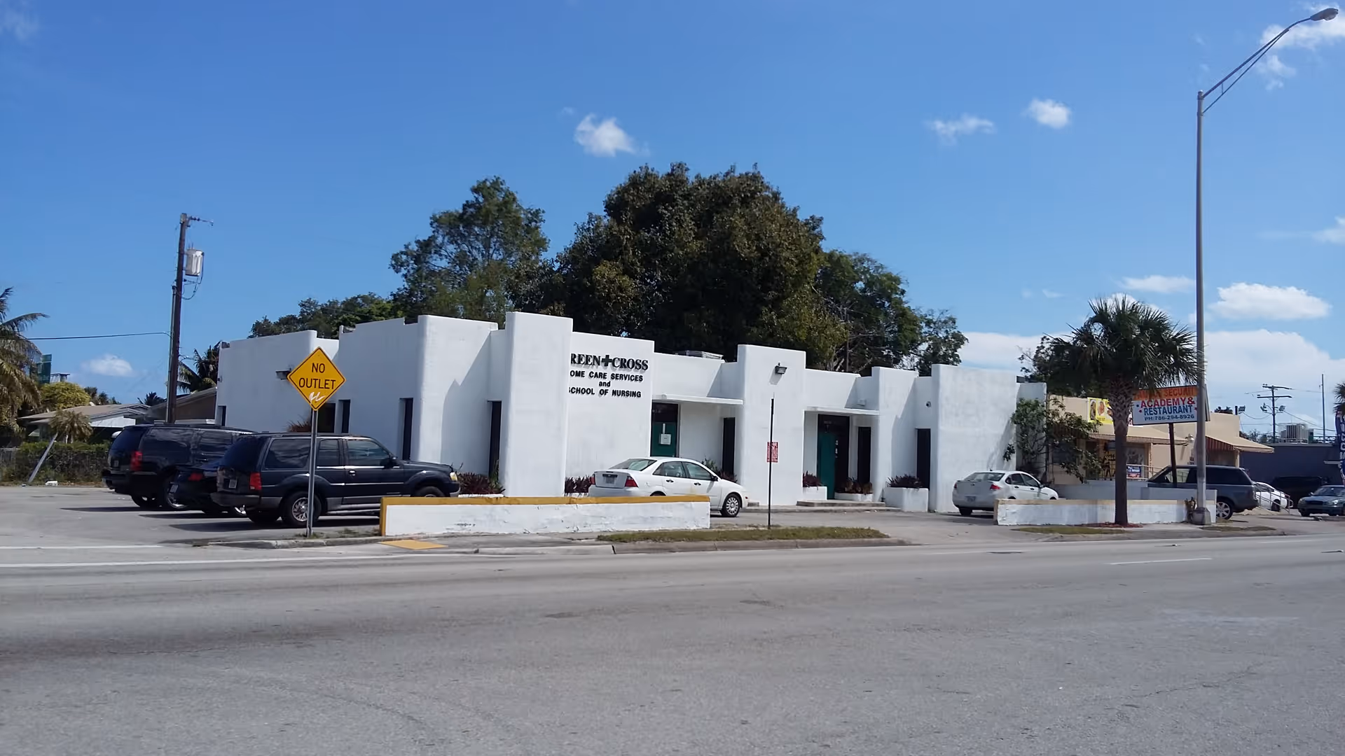 White, modern building labeled Green Cross Home Care Services along a street with parked cars.