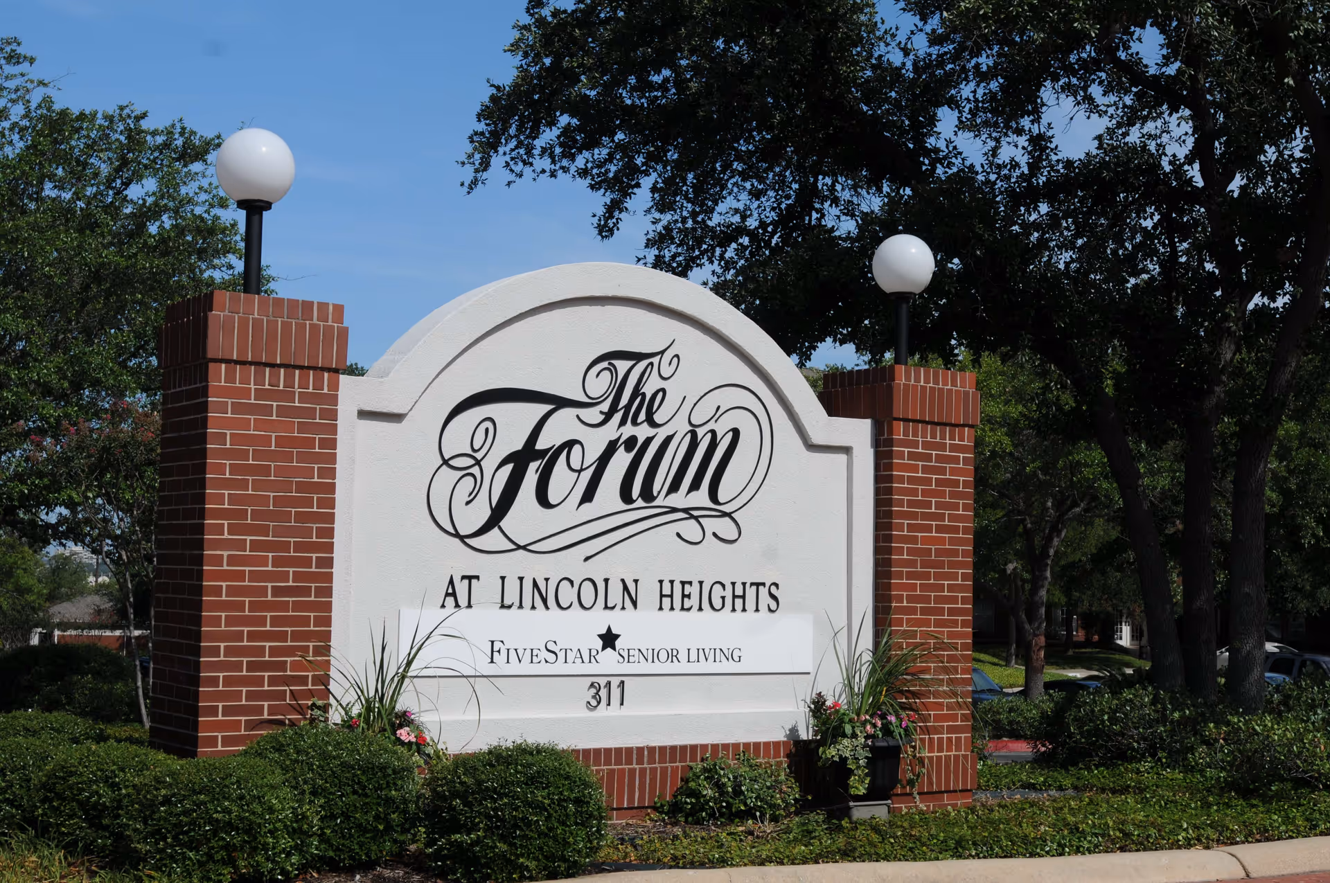Entrance sign reading "The Forum at Lincoln Heights" set between brick pillars with lamps, landscaping, and trees in the background.