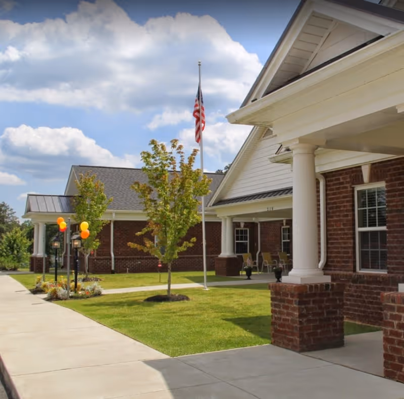 Exterior view of Village Lake Inn of Siler City showing a brick building with white columns and a covered porch. There is a sidewalk, green lawn, small trees, and an American flag on a flagpole under a partly cloudy sky.