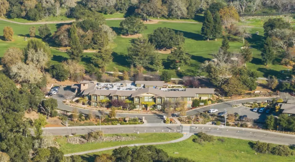 Aerial view of a long senior living building beside a road, surrounded by green lawns and trees.