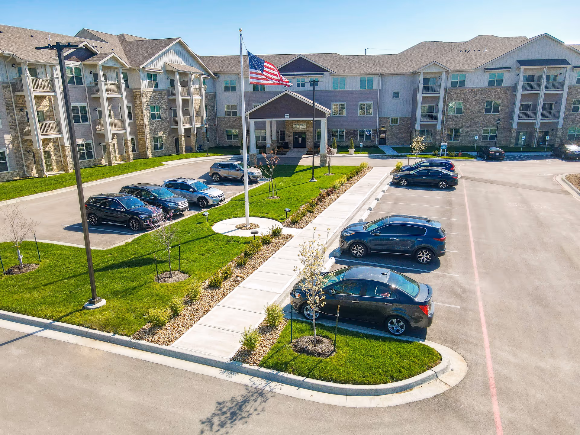 A multi-story senior living facility exterior with a flagpole, parking lot, and landscaped entrance.