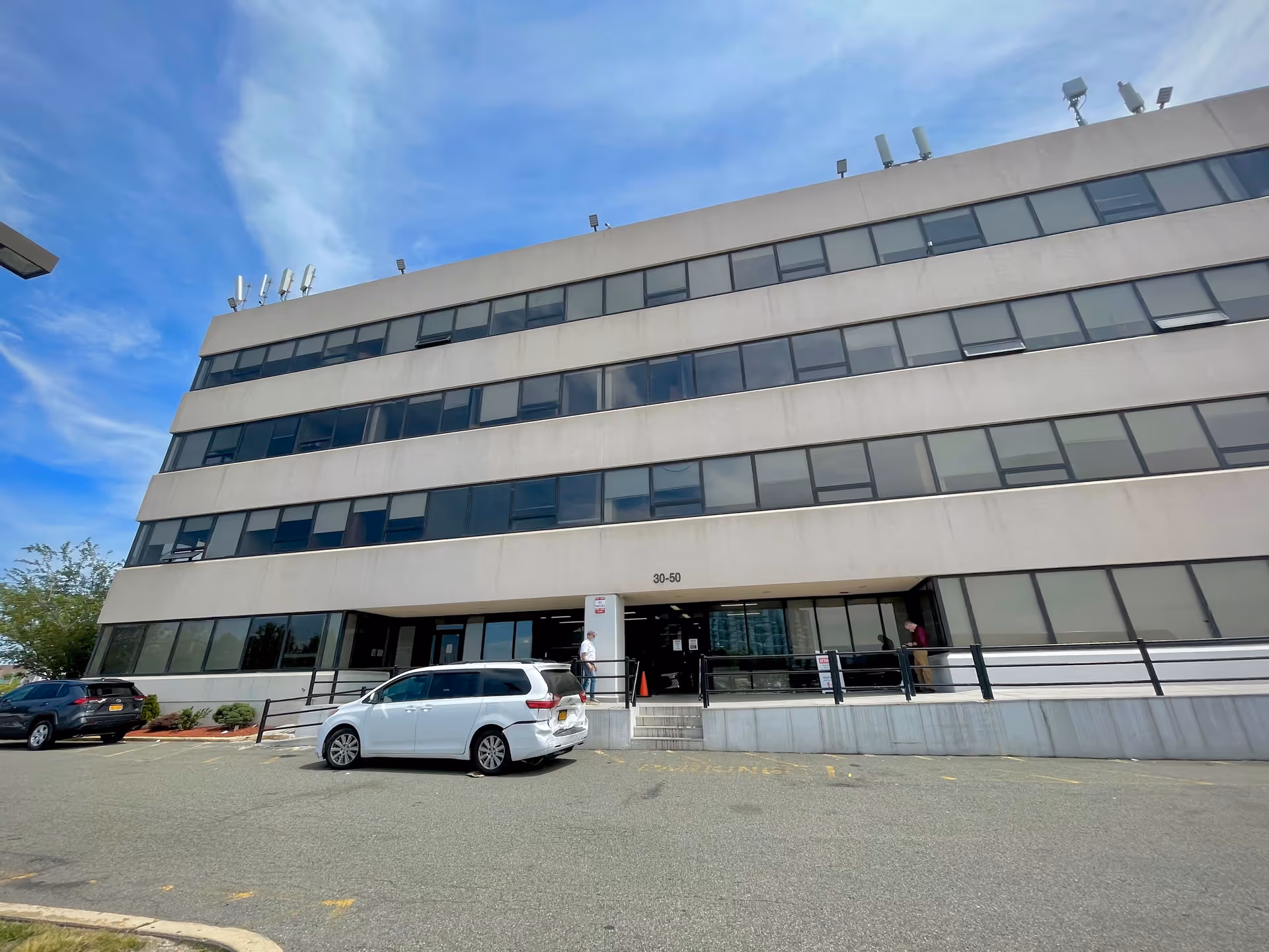 A multi-story beige building facade with a glass entrance and parked cars outside.