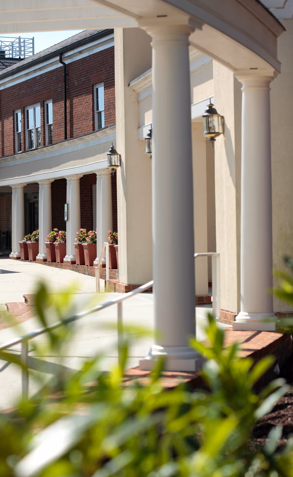 View of the exterior of a building with white columns along a covered walkway, brick walls, potted plants with flowers, and outdoor wall lanterns.