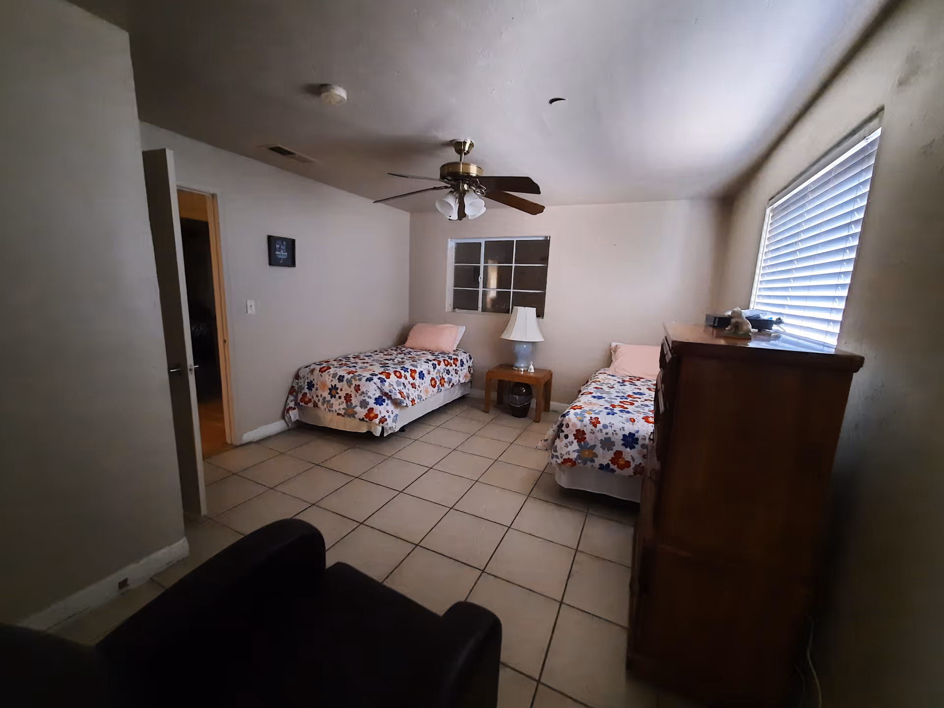A modest bedroom with two single beds covered in floral bedspreads, a wooden dresser, a small wooden nightstand with a lamp, a ceiling fan with lights, and a window with blinds letting in natural light.