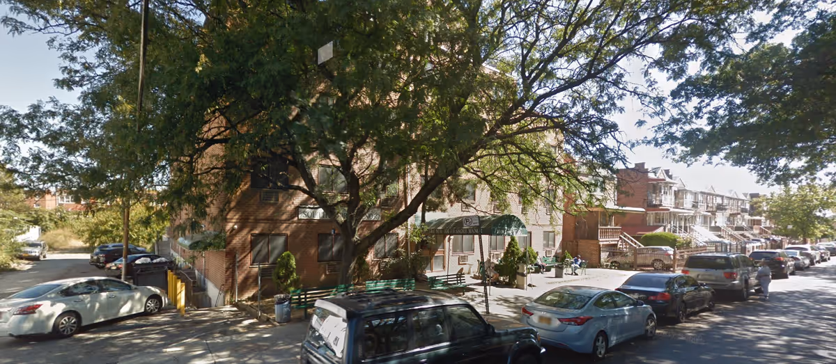 Street view of a brick senior living building, trees, parked cars, and a storefront entrance.