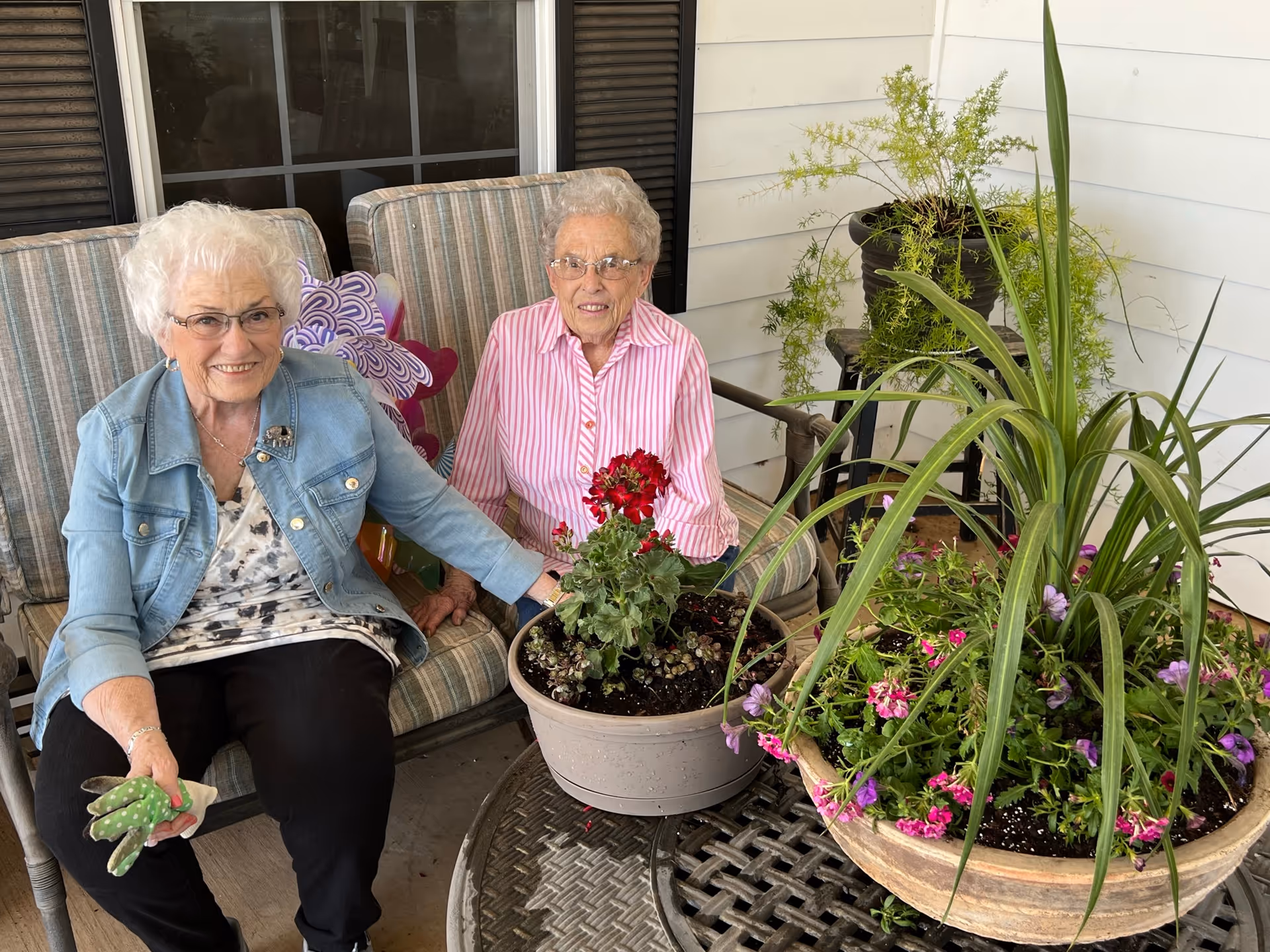 Two elderly women sitting on a cushioned outdoor bench on a porch. One woman is wearing a denim jacket and holding gardening gloves, while the other is wearing a pink and white striped shirt. They are surrounded by potted plants with flowers on a round metal table and nearby surfaces.