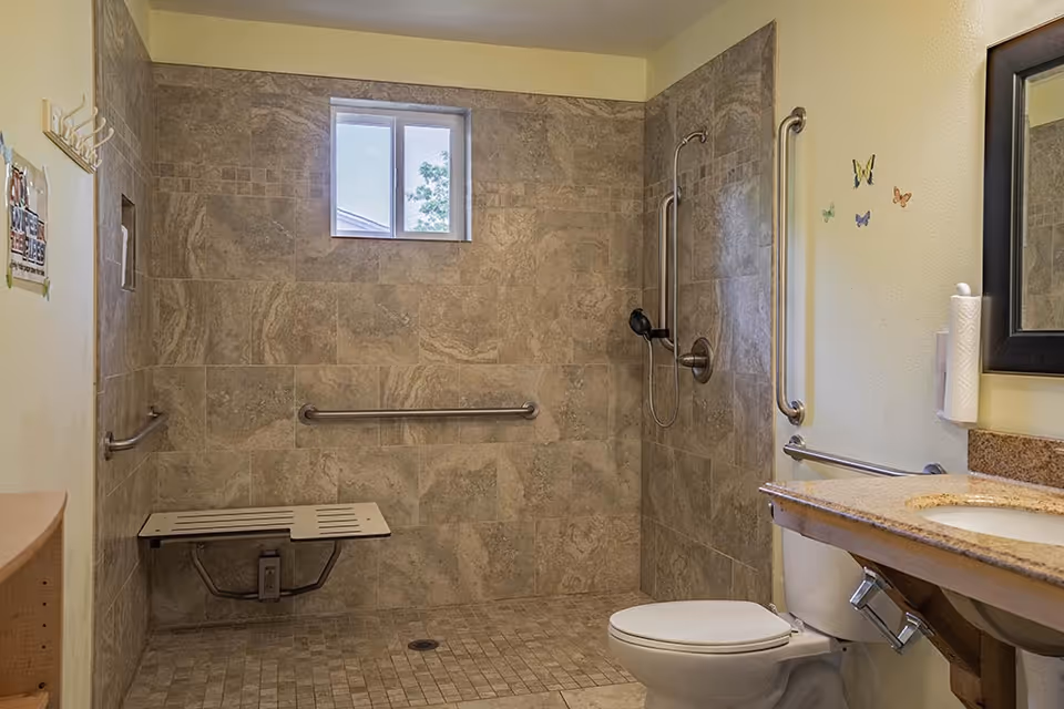 Small window above tiled shower in accessible bathroom with grab bars, bench, toilet, and sink.