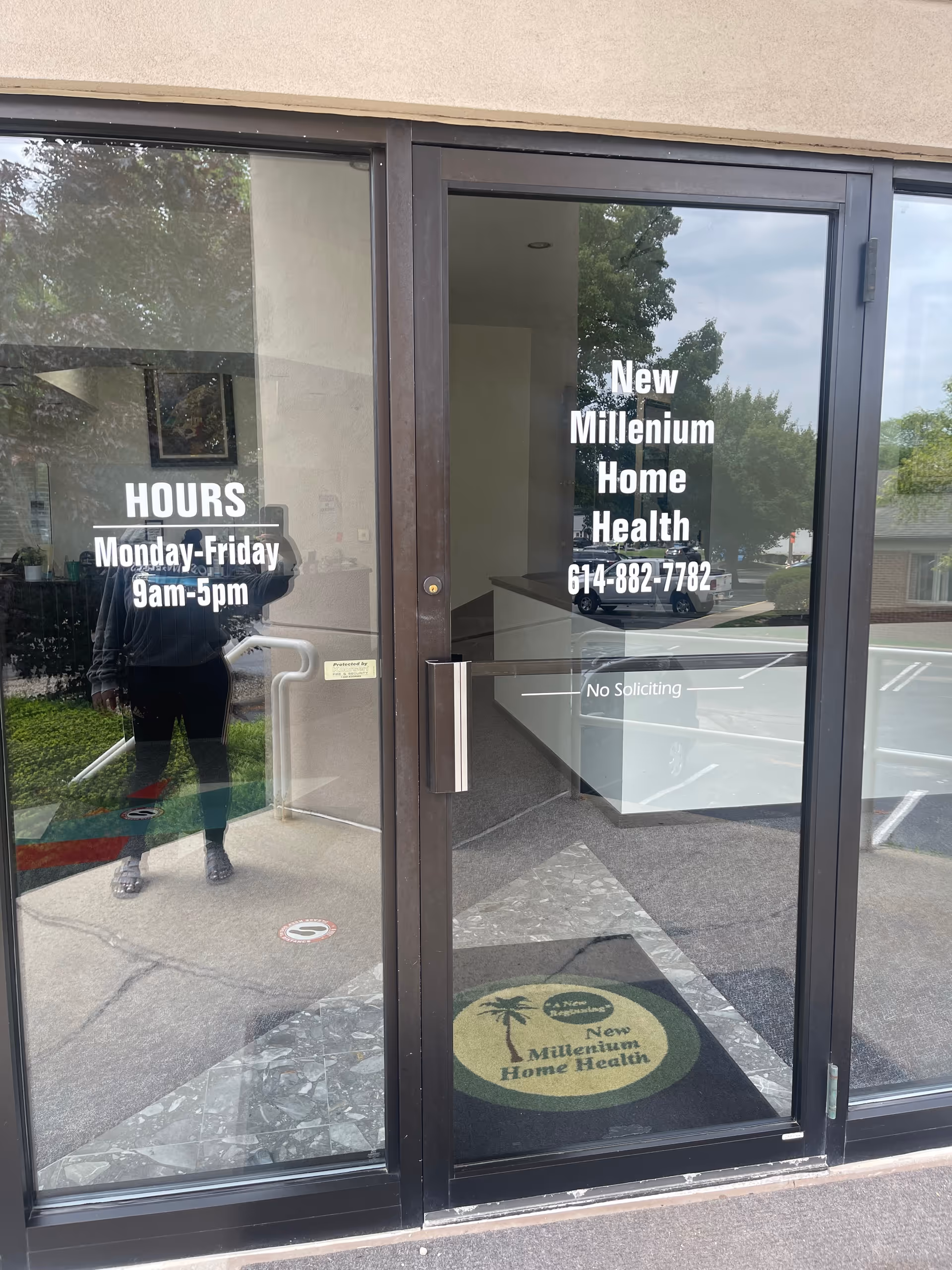 Glass front doors display New Millenium Home Health signage, hours, and a tropical mat outside.