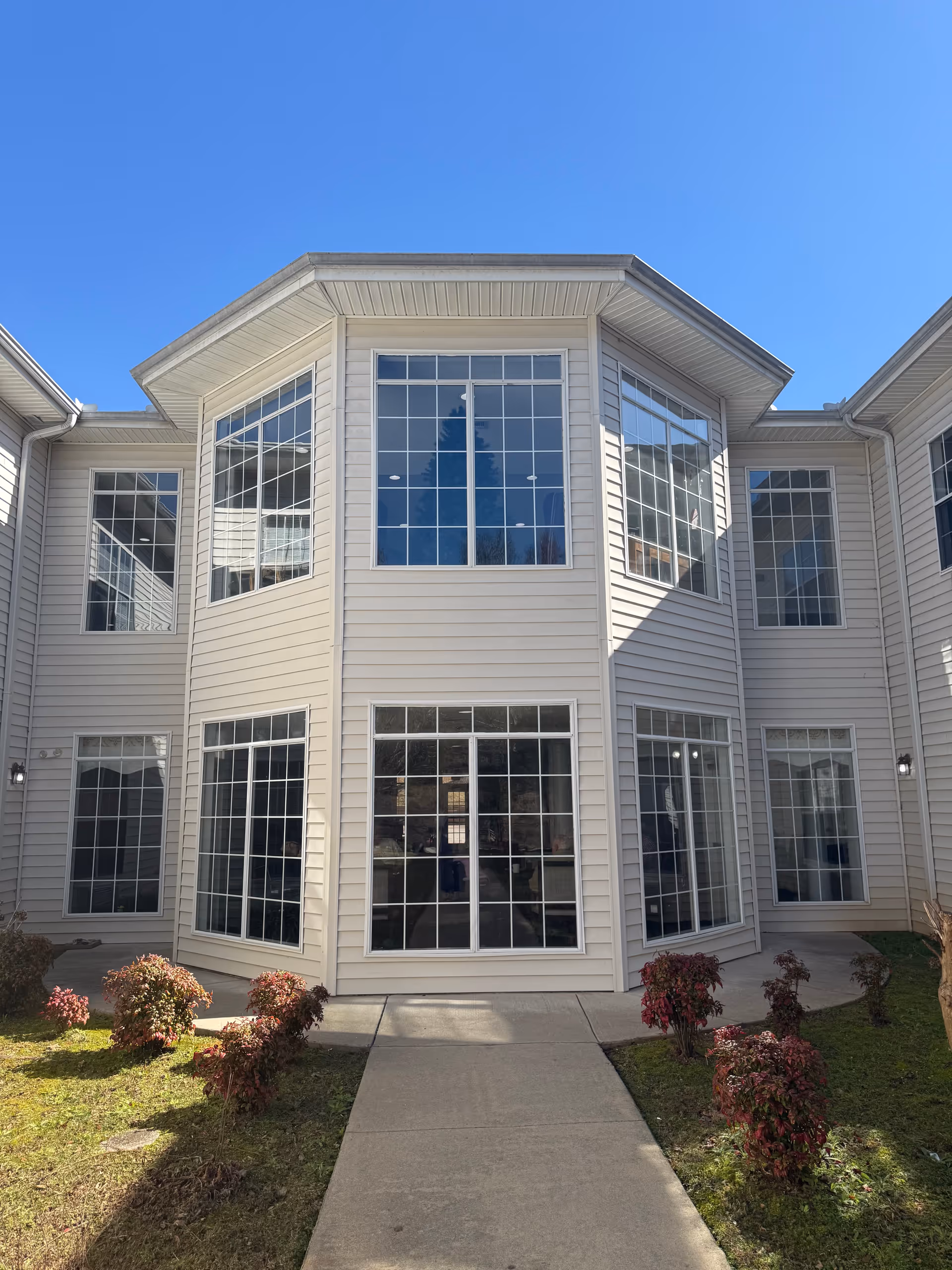 Two-story beige senior living building with large windows, walkway, and shrubs.