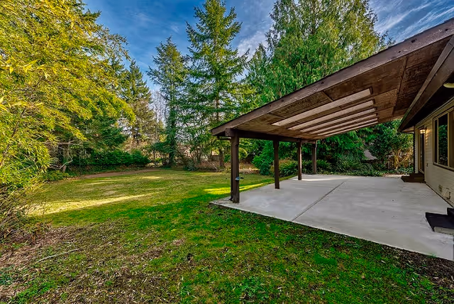 A spacious backyard with a large grassy area surrounded by tall evergreen trees. There is a concrete patio attached to a house, covered by a wooden pergola with beams overhead. The sky is partly cloudy with blue patches visible.